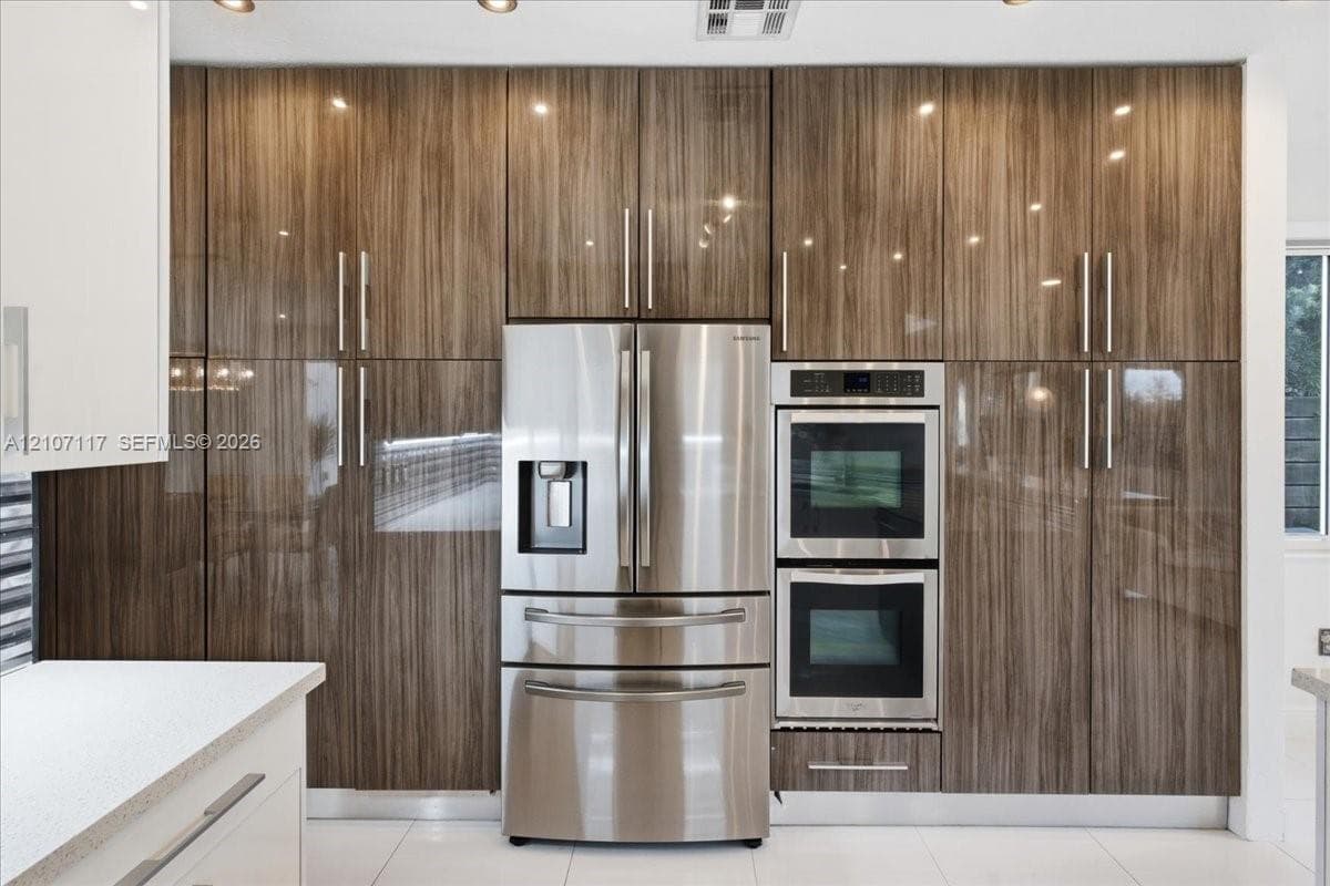 Full-height high-gloss wood-finish cabinetry, stainless steel French door refrigerator, built-in double wall oven, and light-toned solid surface countertops