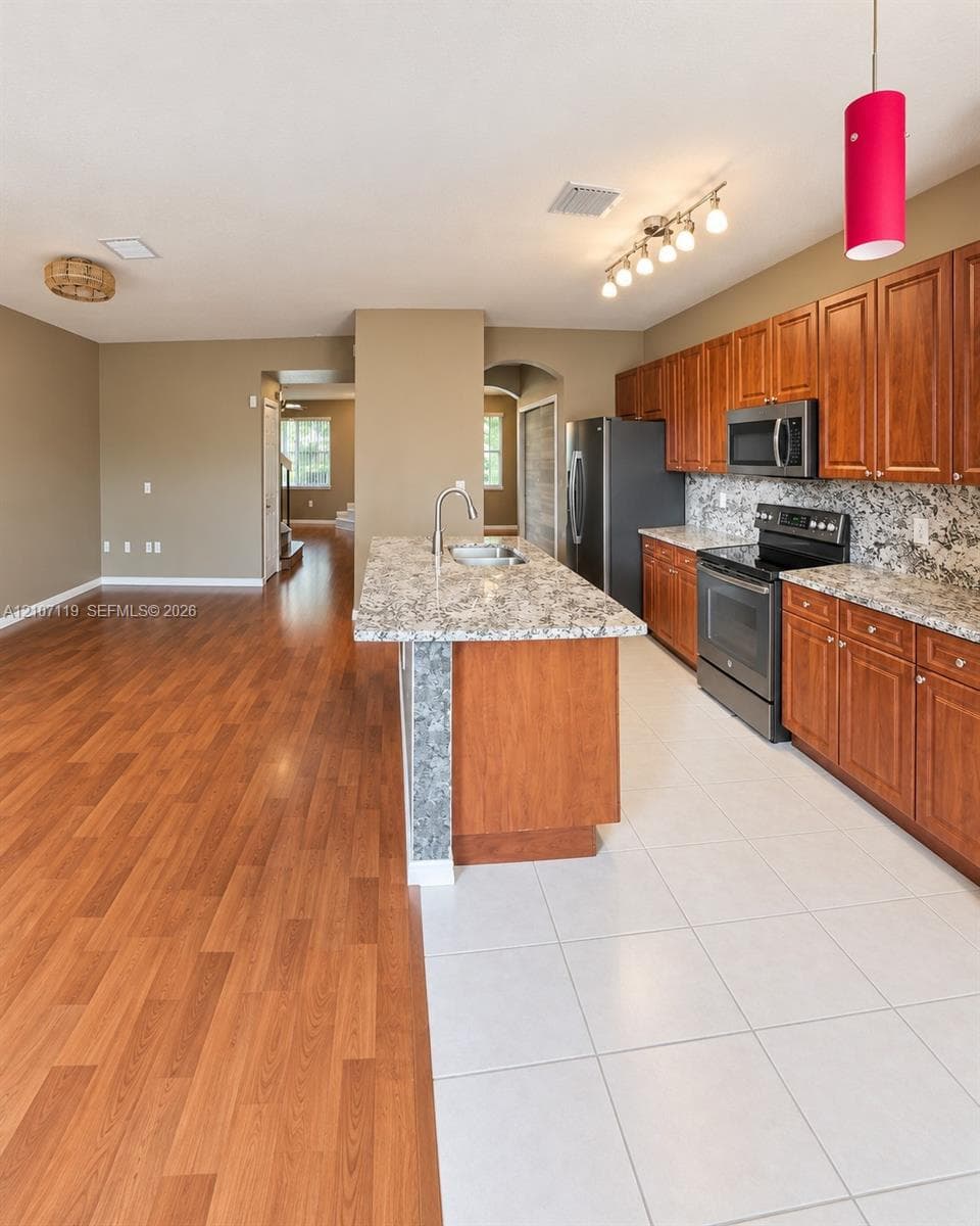 Kitchen featuring wood-finish cabinetry, stone-look countertops, an island with an undermount sink, ceramic tile flooring, and integrated stainless steel appliances