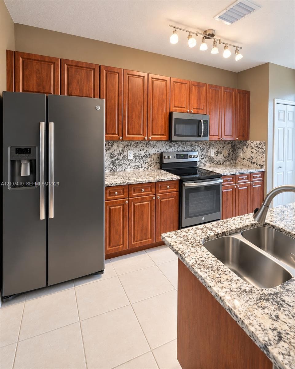Kitchen featuring wood cabinetry, stone-finish countertops, stainless steel appliances, multi-basin sink with pull-down faucet, and track lighting