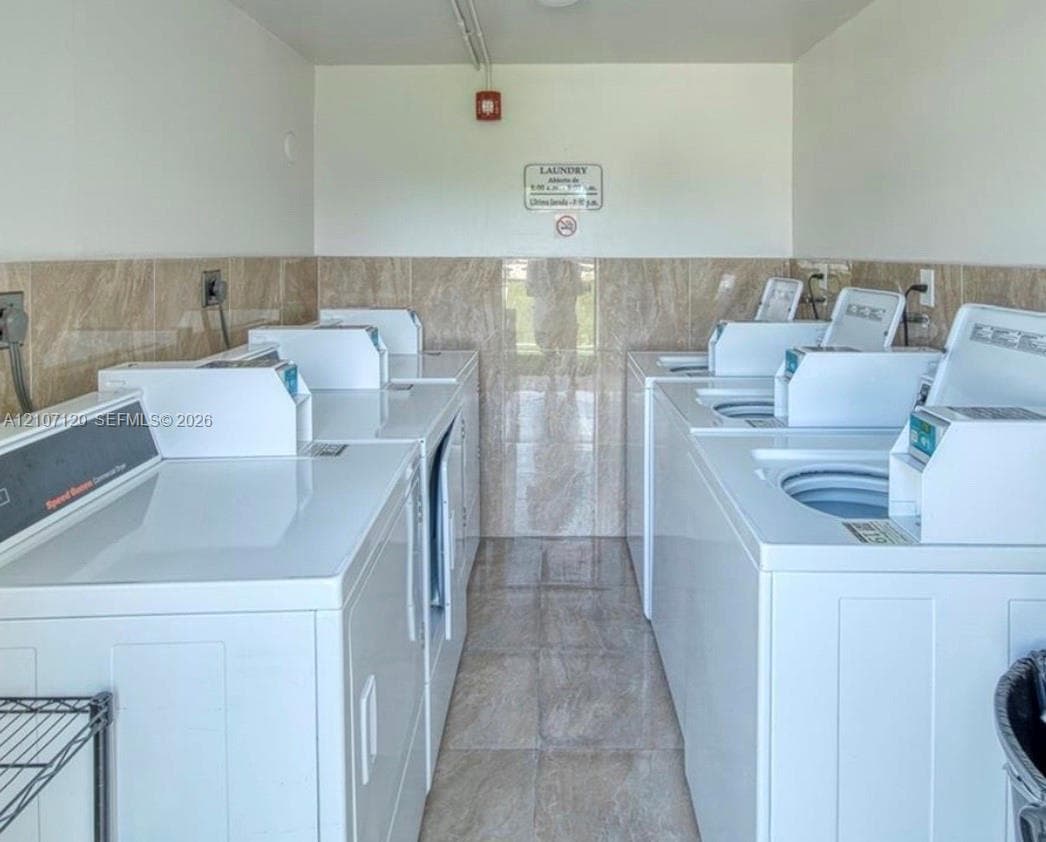 Laundry facility featuring multiple commercial-grade top-load washing machines and dryers, ceramic tile flooring, and half-height ceramic wall tiling