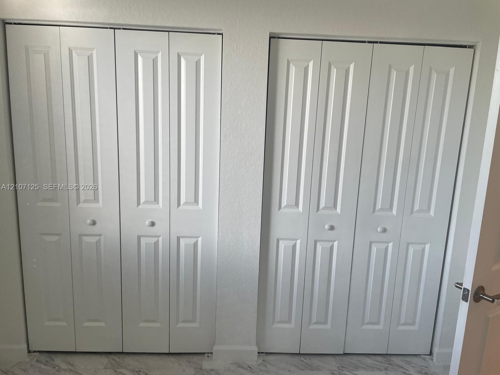 Dual sets of white bi-fold closet doors featuring recessed panel detailing and round knobs, complemented by neutral-toned stone-finish flooring