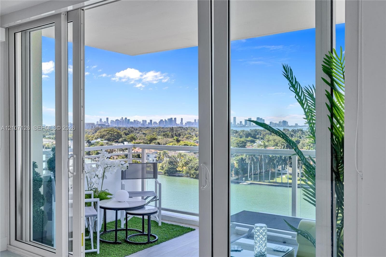 Full-height glass sliding doors opening to a covered balcony with a white metal railing, overlooking an expansive waterway and metropolitan skyline
