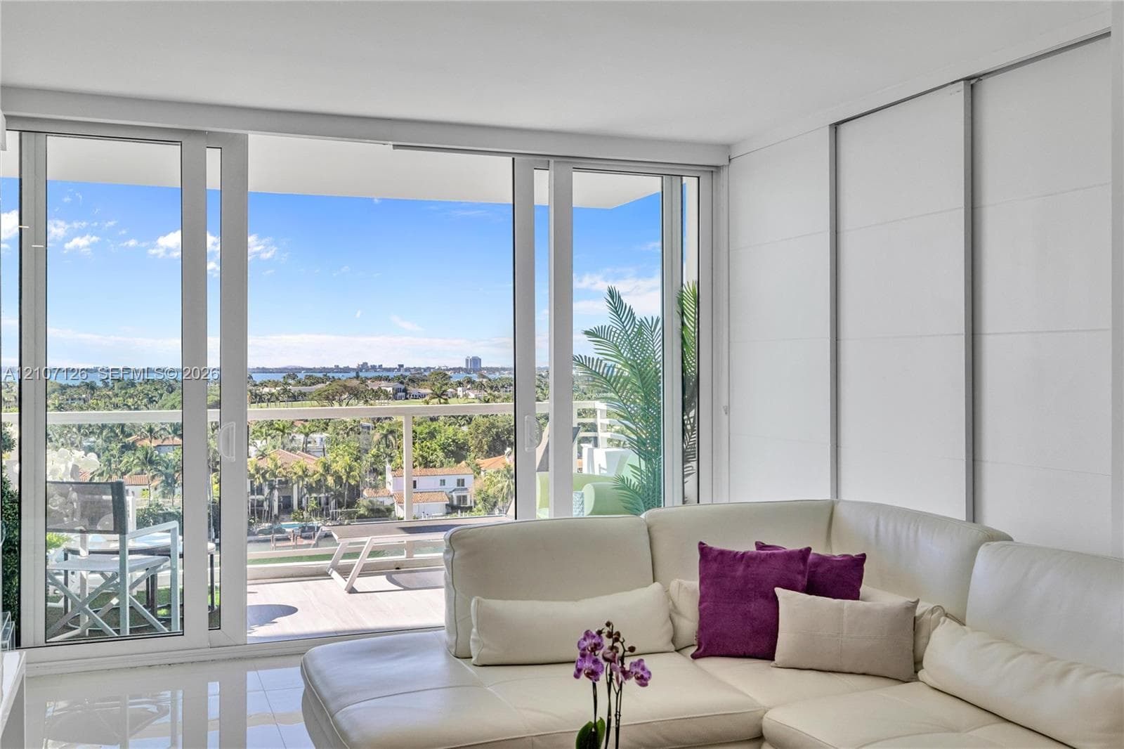 Floor-to-ceiling sliding glass doors opening to a covered balcony with a glass railing, white polished flooring, and expansive exterior water and landscape scenery