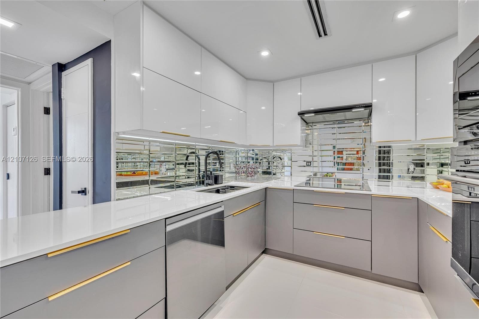 Modern kitchen featuring high-gloss white upper cabinetry, grey flat-panel lower cabinetry, gold-tone hardware, white stone countertops, and a mirrored tile backsplash
