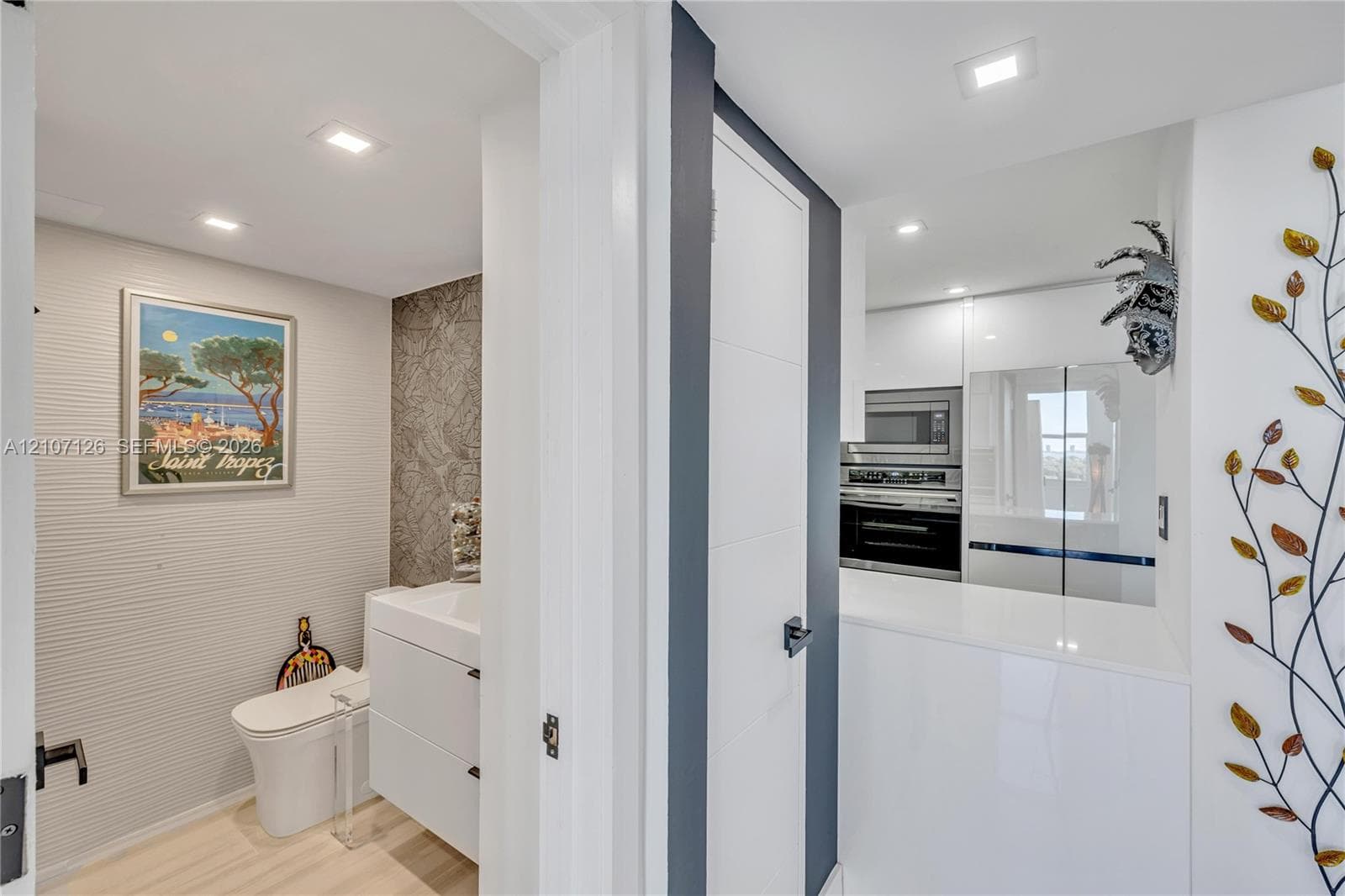Modern bathroom featuring a sleek wall-hung vanity, textured wall surfacing, and recessed lighting, paired with a kitchen area offering crisp white cabinetry, built-in stainless steel wall oven, and a solid-surface countertop