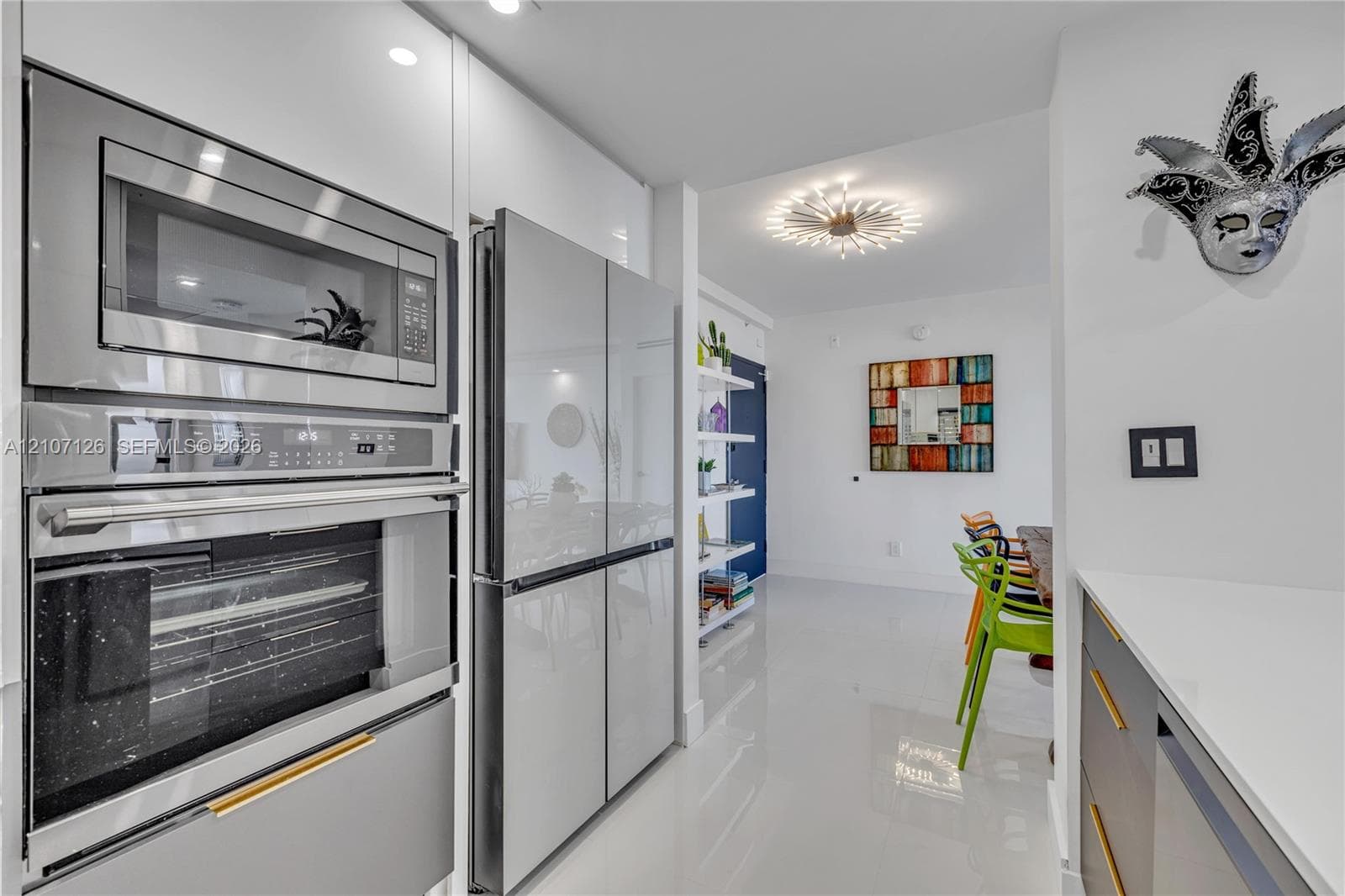 Modern kitchen featuring glossy white cabinetry, stainless steel wall-mounted oven and microwave combination, four-door refrigerator with matching finish, stone-surface countertops, and sleek gold-toned hardware