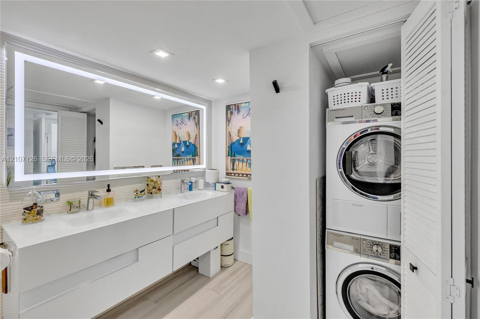 Floating dual-sink vanity with a solid-surface countertop, full-width LED-backlit mirror, wood-finish flooring, and integrated stackable laundry unit behind louvered closet doors