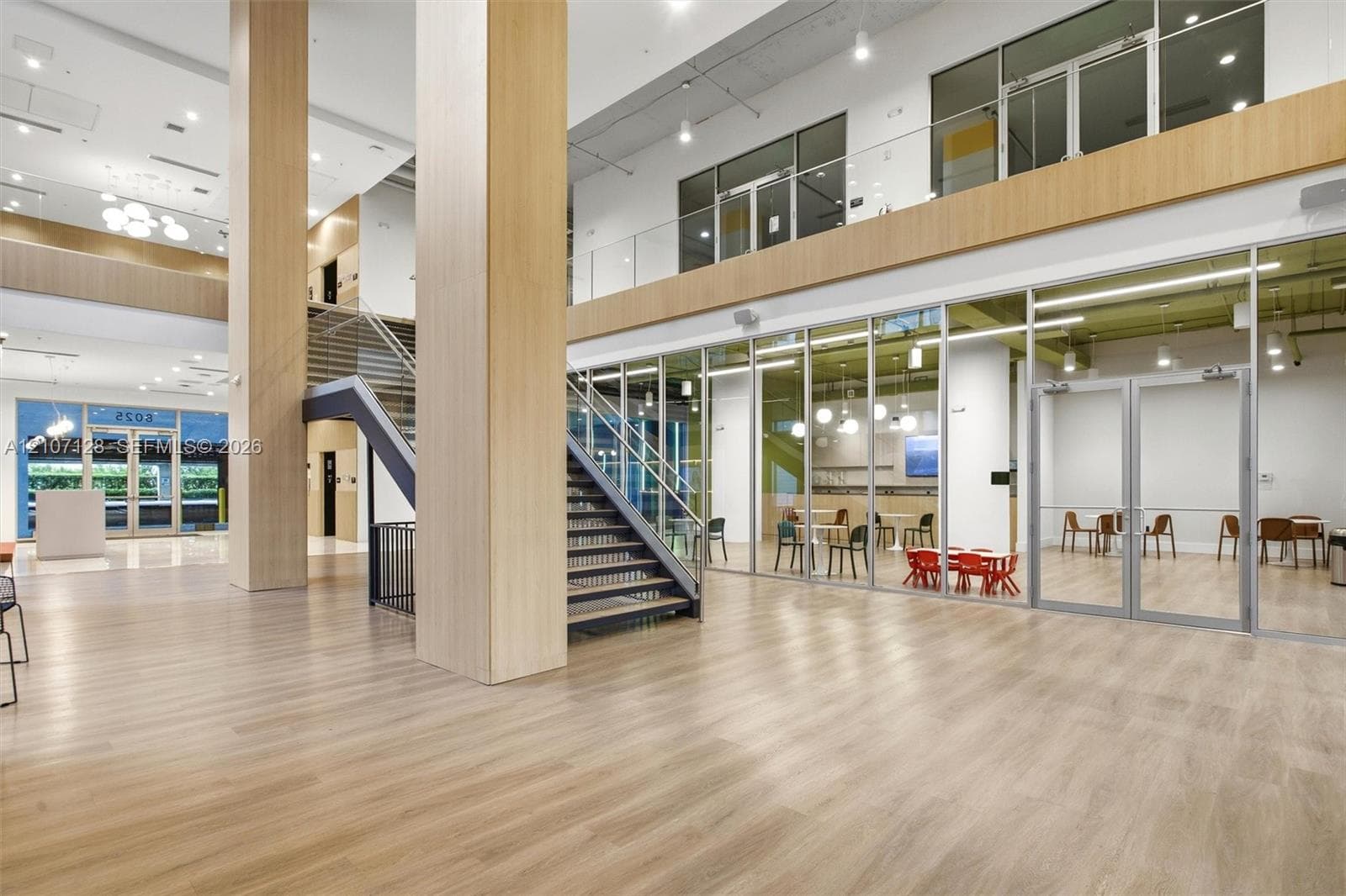 Atrium featuring a double-height ceiling, wood-finish flooring, floor-to-ceiling glass partitions, a prominent central staircase with metal railings, and dual-level mezzanine construction