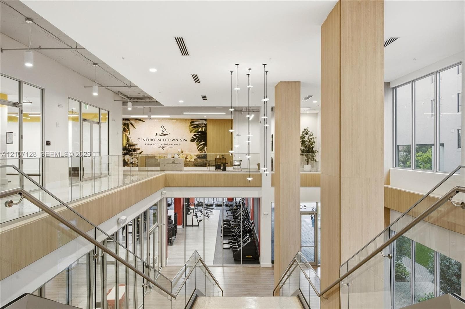 Multi-level interior space featuring wood-finish paneling, floor-to-ceiling glass partitions, a glass-panel staircase railing, large windows, and a multi-pendant lighting fixture