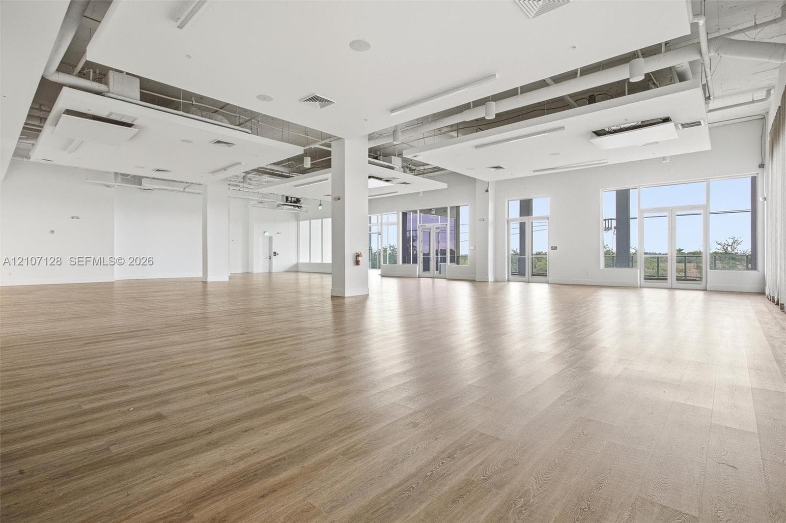 Expansive open-concept interior featuring wood-finish flooring, suspended ceiling architectural panels, structural columns, and floor-to-ceiling windows with glass-panel doors