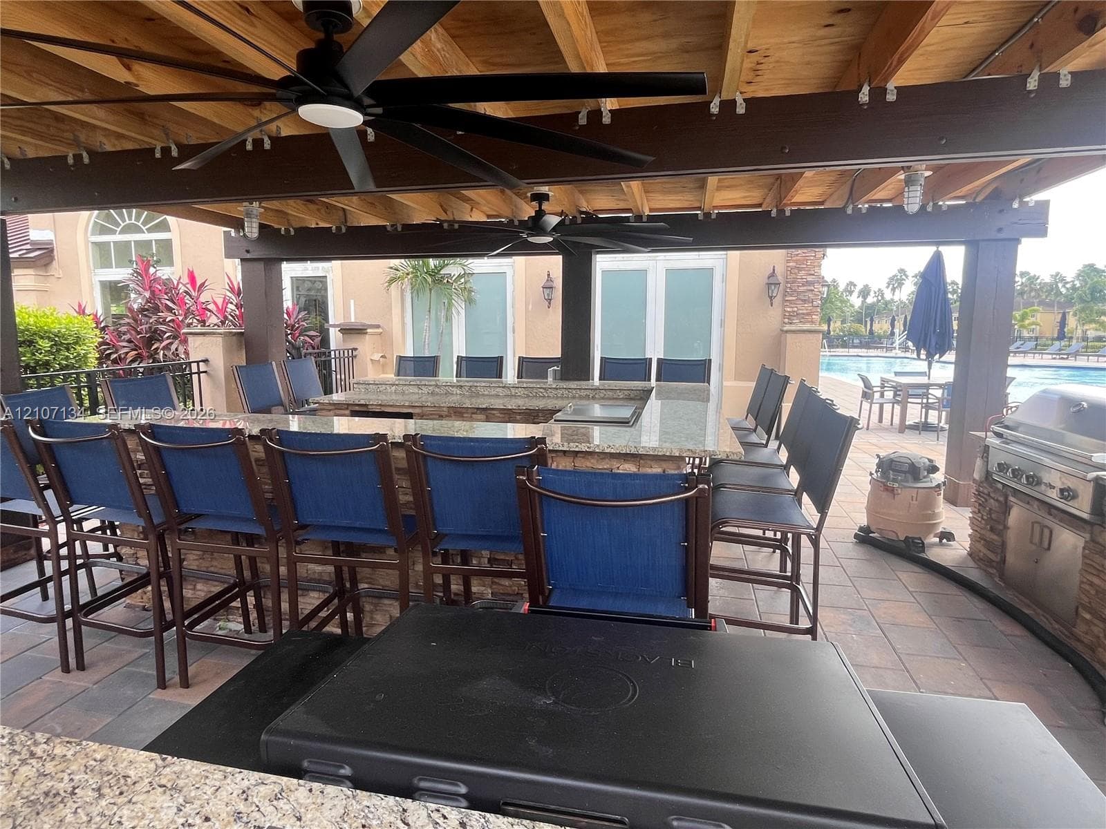 Covered outdoor kitchen featuring stone masonry, a stone-topped bar, a built-in stainless steel grill, and dual ceiling fans under a timber-framed pergola