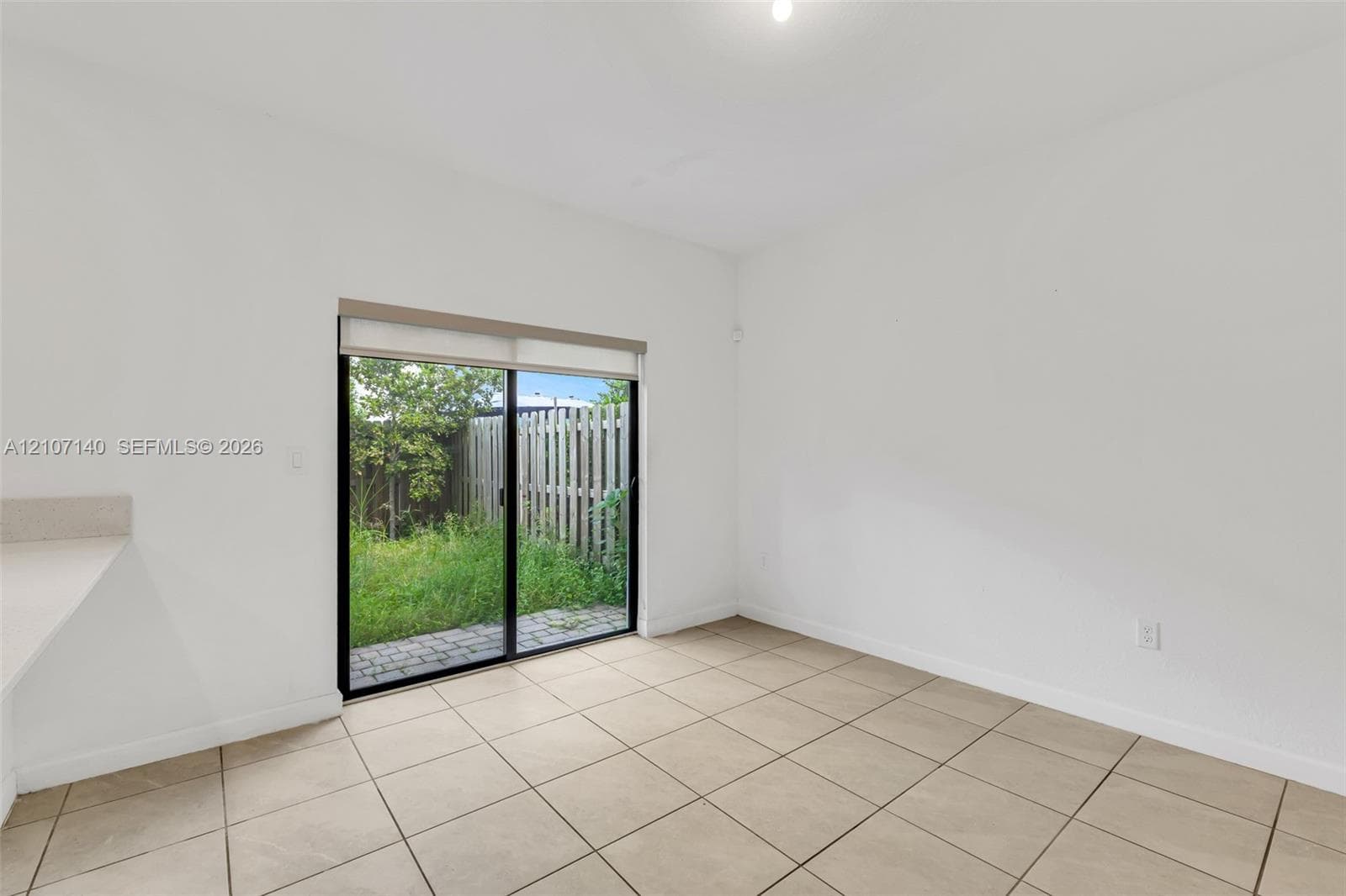 Interior featuring ceramic tile flooring, neutral walls, and a sliding glass door with a roller shade, leading to a fenced outdoor area with paver hardscaping