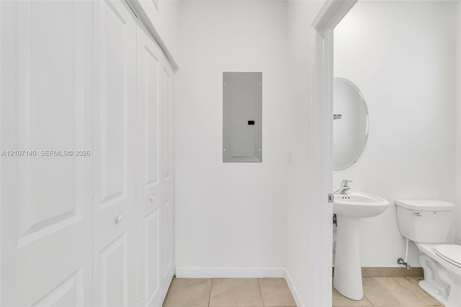 White bi-fold doors, porcelain pedestal sink, oval wall-mounted mirror, ceramic tile flooring, and integrated electrical service panel