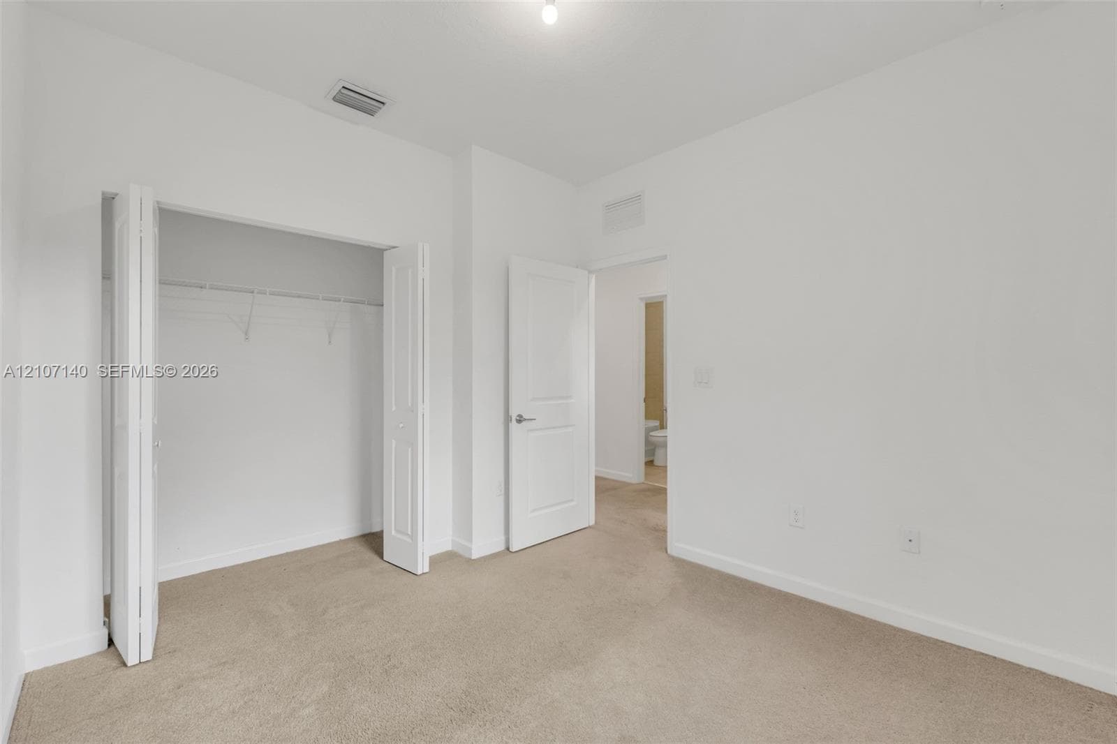 Neutral-toned interior space featuring beige carpeting, white walls, a multi-panel door, and a closet with bifold doors
