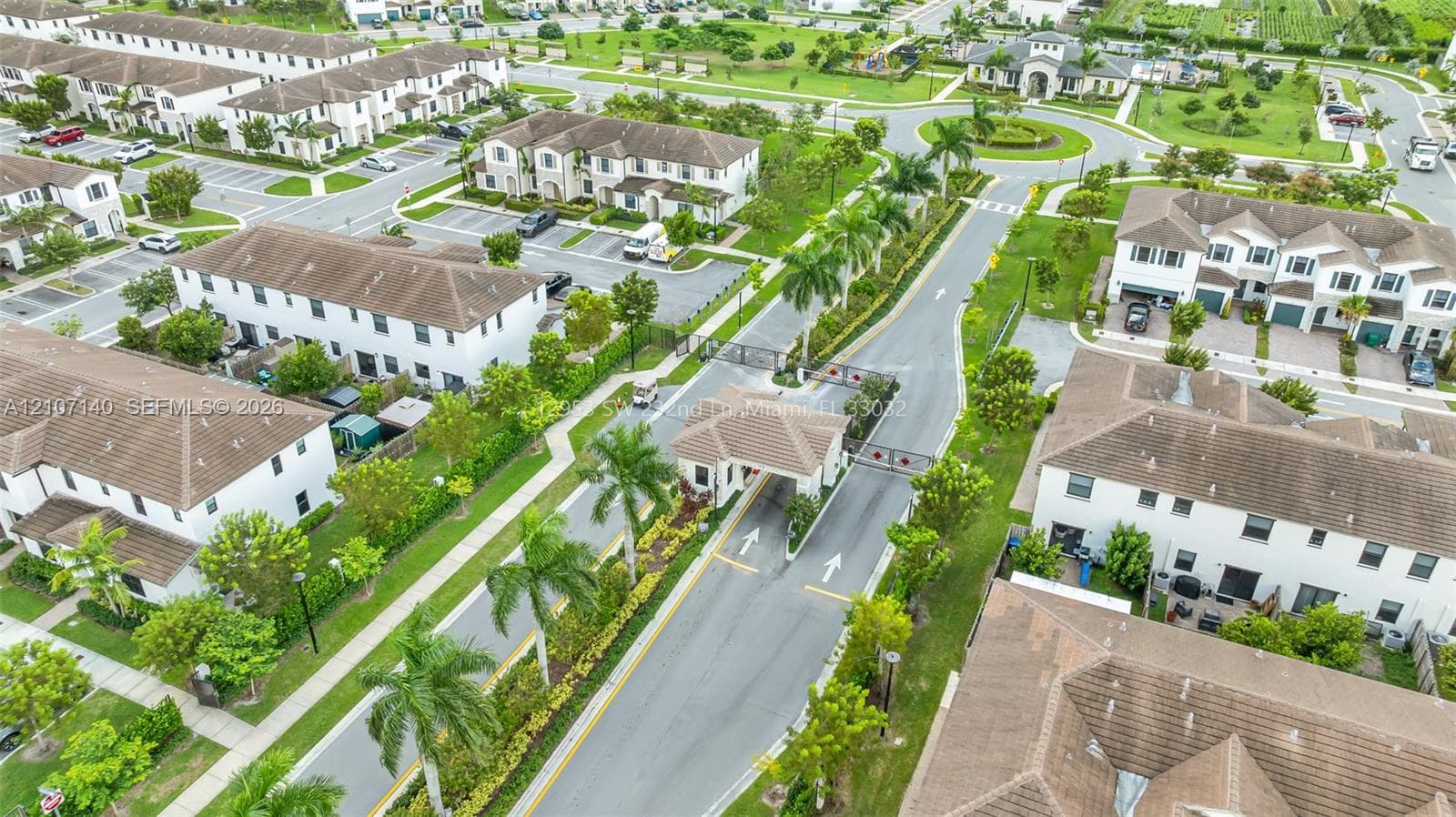 Gated entrance with a security checkpoint, multi-unit residential buildings with tile roofing, asphalt thoroughfares, and manicured landscaping featuring mature palm trees