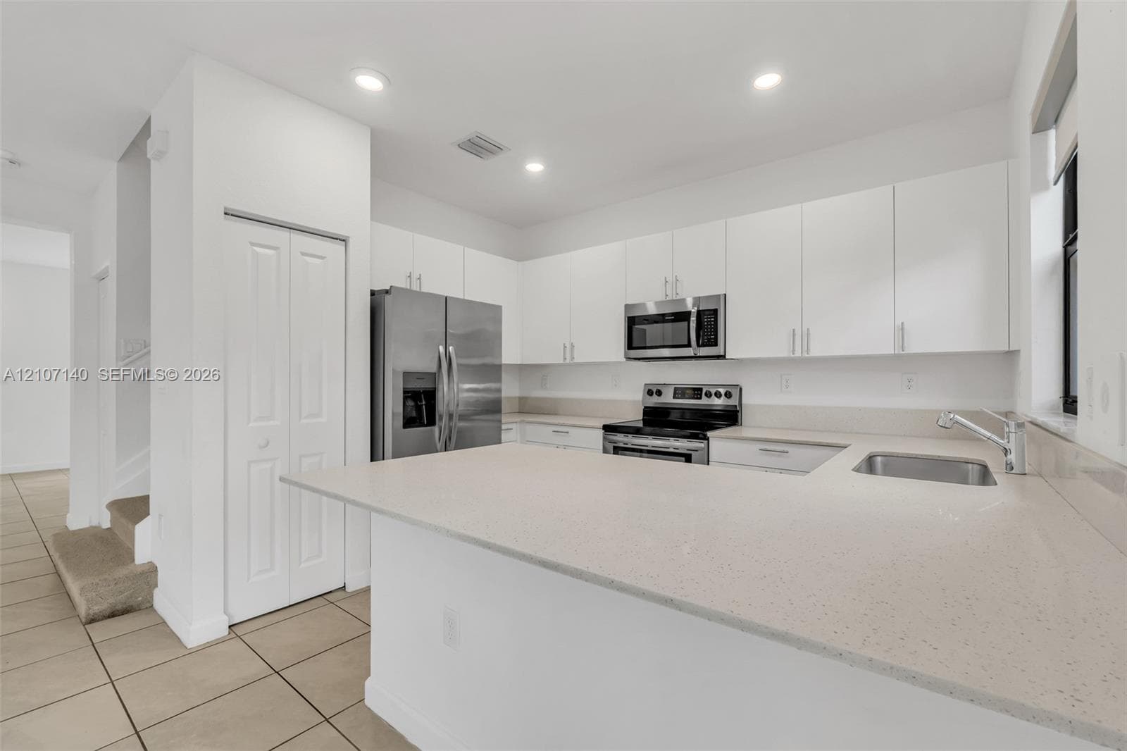 Kitchen featuring white flat-panel cabinetry, light-toned stone-finish countertops, stainless steel appliances, a peninsula with an integrated sink, and tile flooring