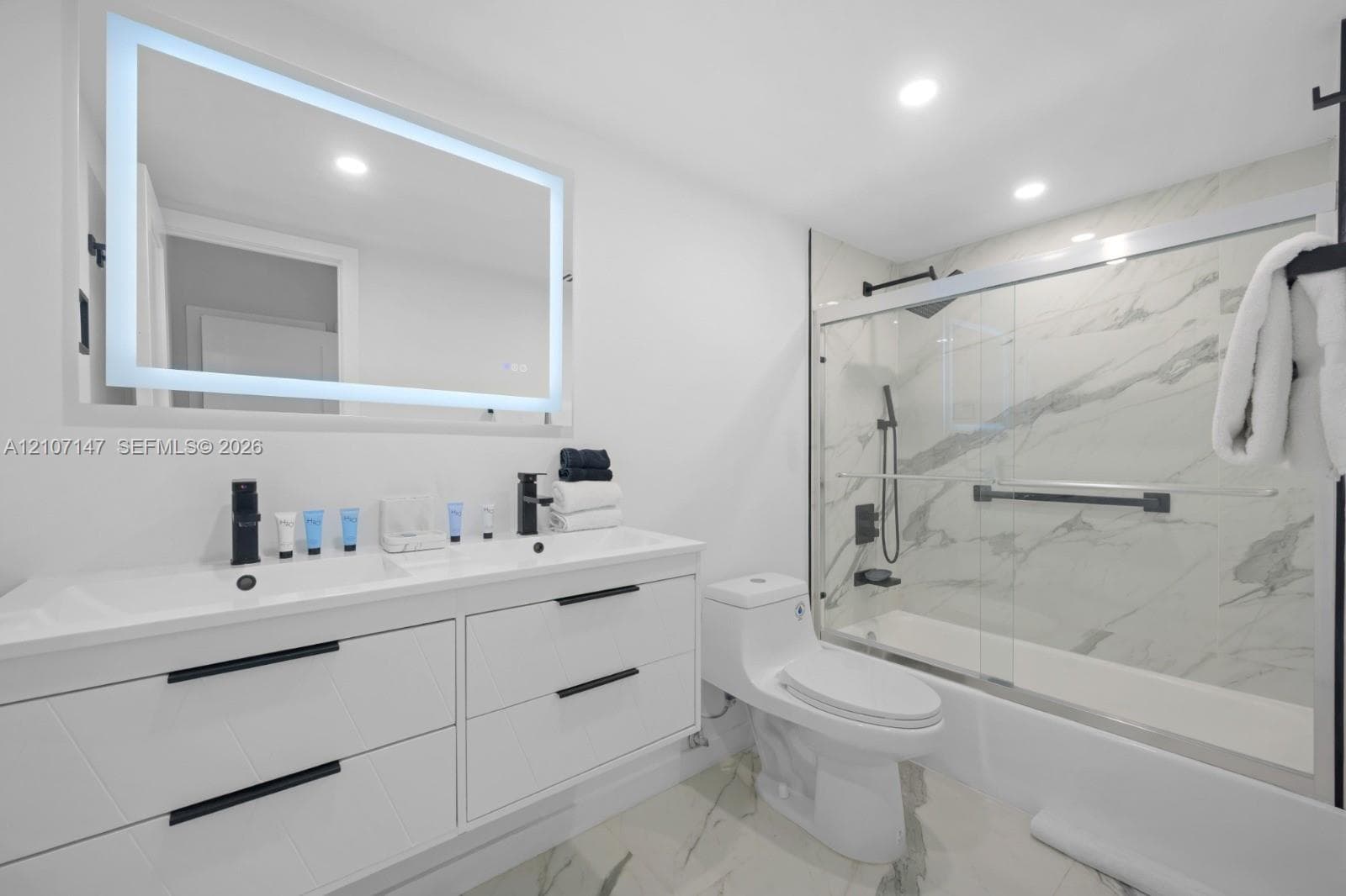 Modern bathroom featuring a double-sink vanity with matte black hardware, backlit LED mirror, recessed ceiling lighting, and a bathtub-shower combination enclosed by a glass door with large-format stone-patterned tile surround