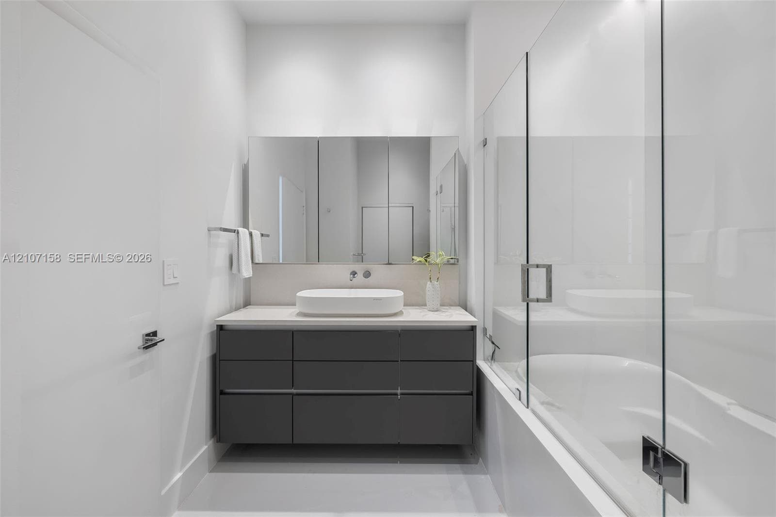 Modern floating vanity with a rectangular vessel sink, solid surface countertop, matte finish cabinetry, expansive mirrored medicine cabinet, and glass-enclosed tub with chrome hardware