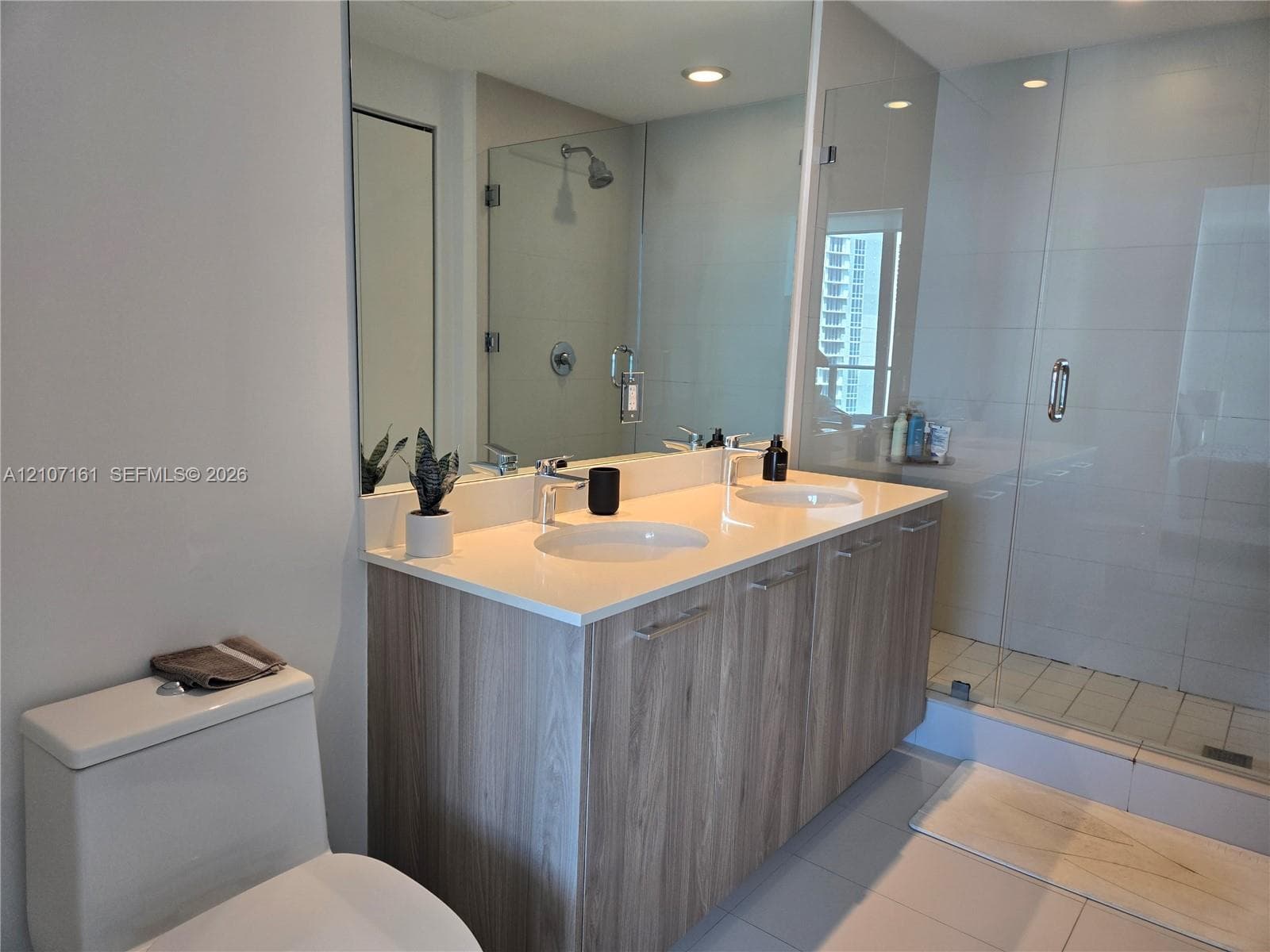 Dual-sink vanity with light wood-finish cabinetry, solid-surface countertop, full-width mirror, and a glass-enclosed shower with tiled walls