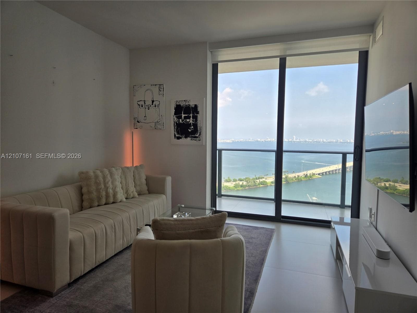 Floor-to-ceiling glass sliding doors opening to a balcony with expansive water and causeway scenery, complemented by large-format tile flooring