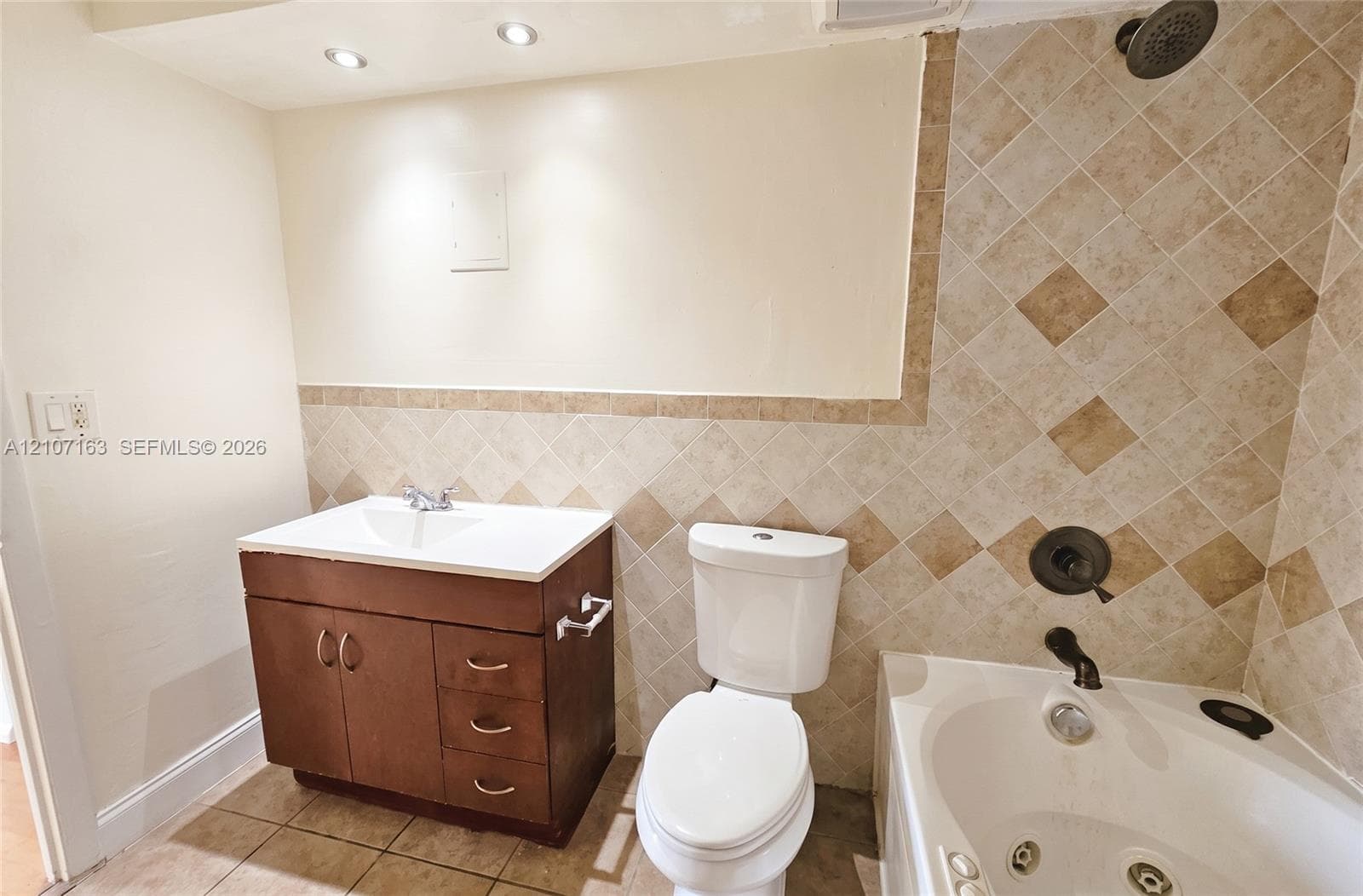 Bathroom featuring a wood-finish vanity with an integrated basin, tile wainscoting with decorative inlay, recessed ceiling lighting, and a jetted soaking tub with a multi-piece shower fixture