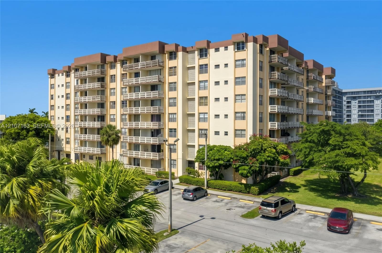 Multi-story residential building featuring private balconies, masonry exterior, concrete parking surface, and established landscaping