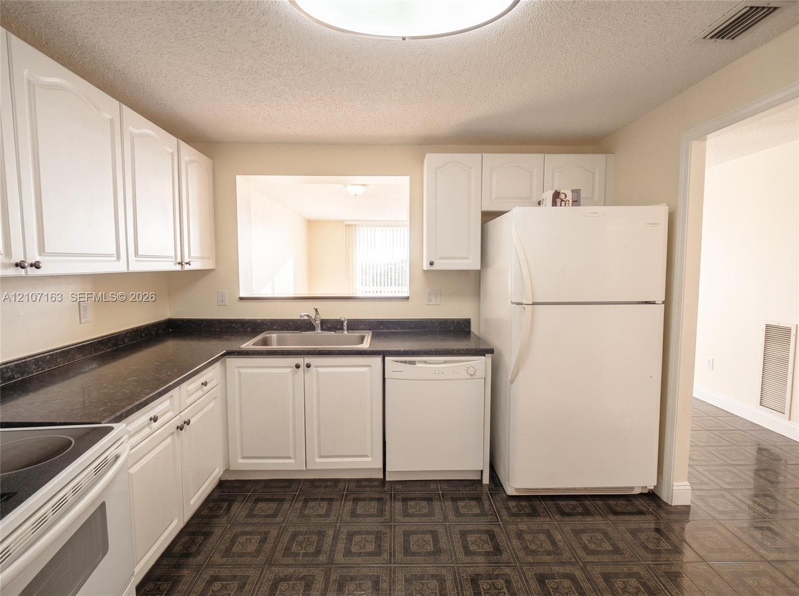 Kitchen featuring white cabinetry, dark countertops, a stainless steel sink with a chrome faucet, white appliances, and patterned tile flooring