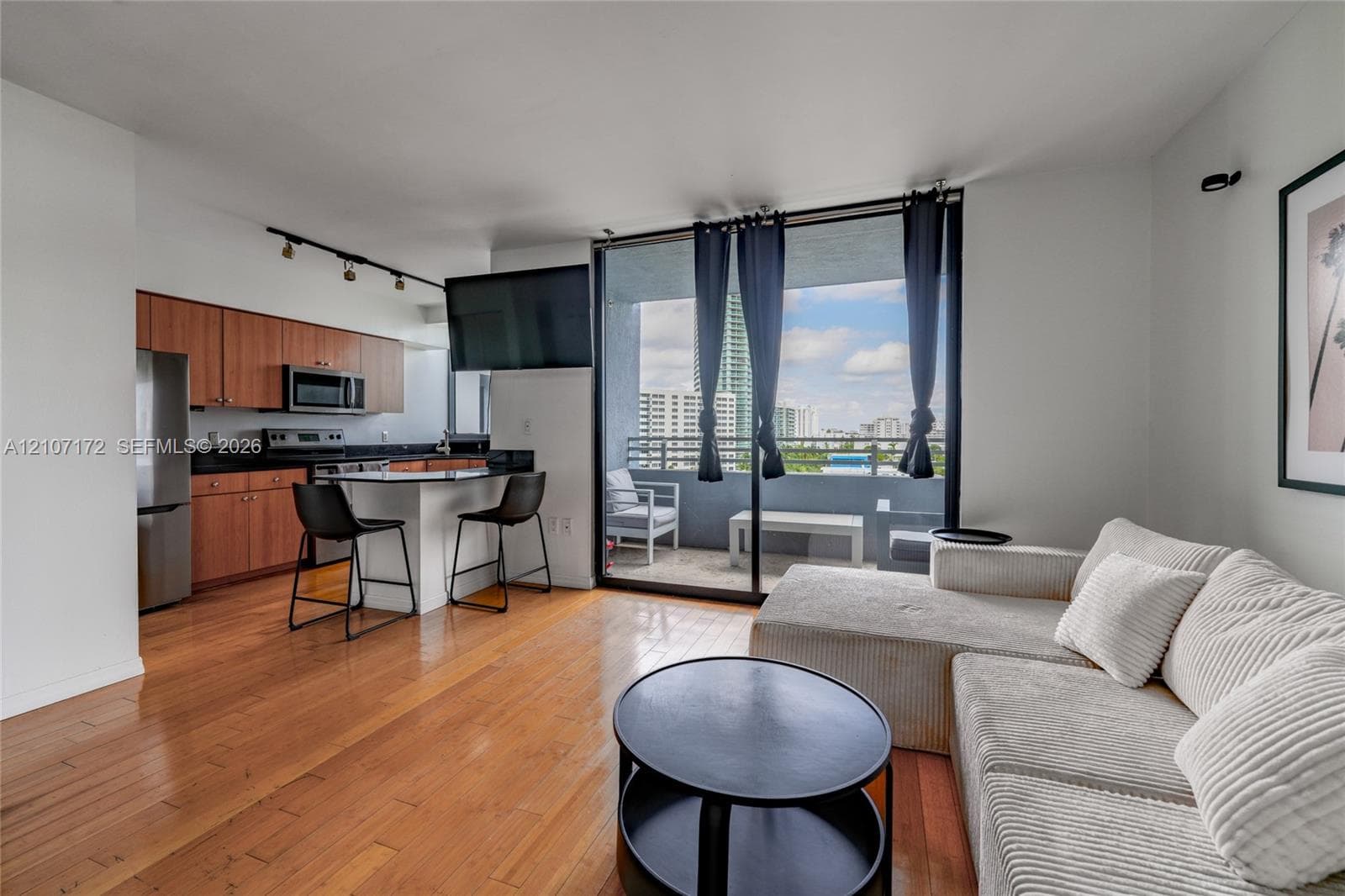 Open-concept kitchen featuring wood-finish cabinetry, stone countertops, and integrated stainless steel appliances, complemented by wood-finish flooring and floor-to-ceiling glass sliding doors leading to a private balcony