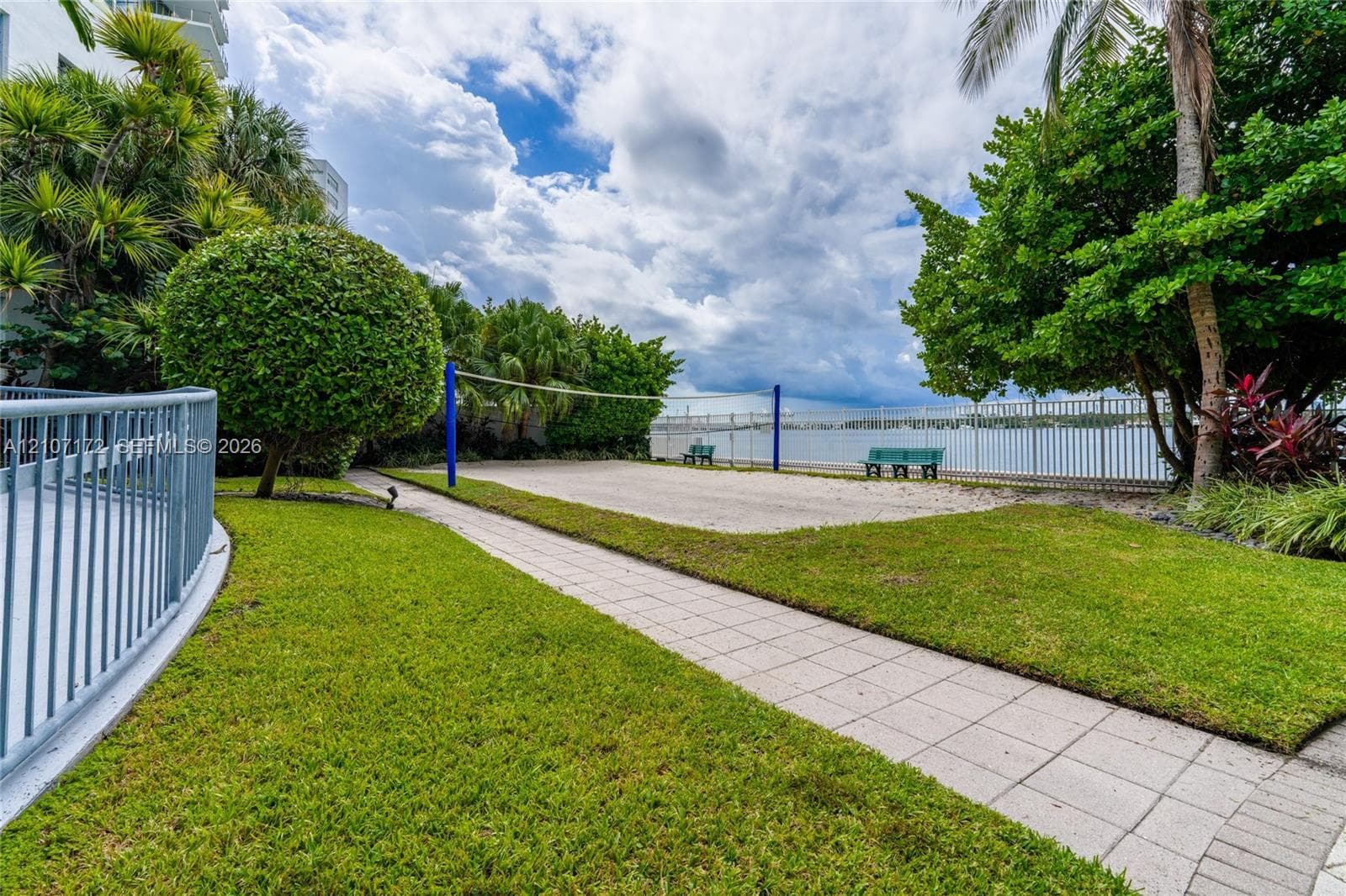 Sand volleyball court featuring a net, bordering a paved stone walkway, manicured lawn, and water frontage