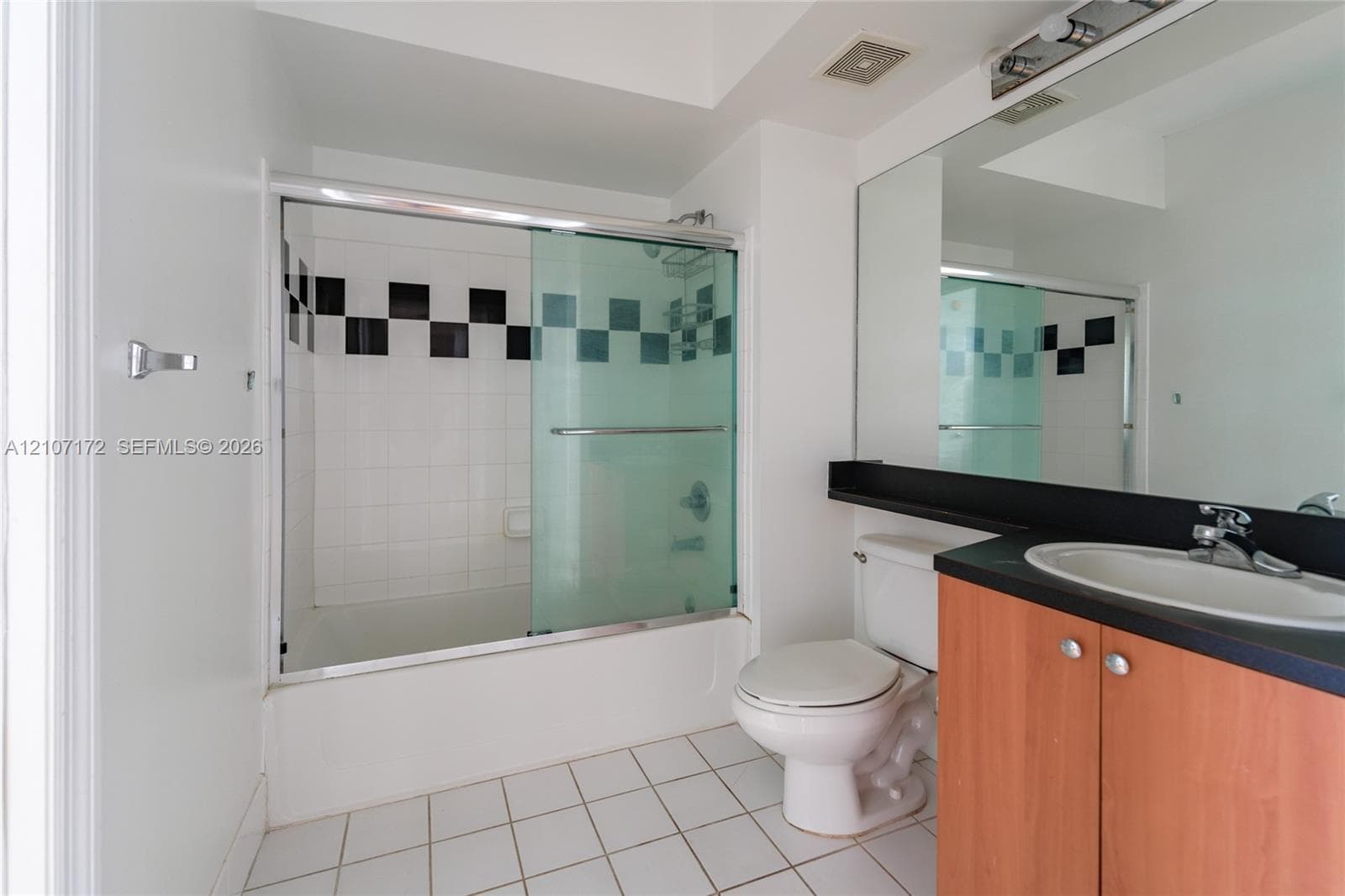 Bathroom featuring a vanity with wood-finish cabinetry, a black countertop with an integrated oval sink, a wall-to-wall mirror, and a shower-tub combination with sliding glass doors and decorative tile inlay