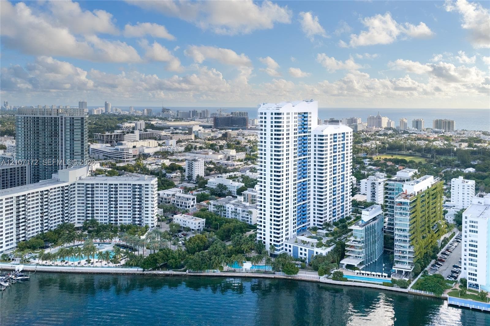 Aerial perspective of a high-rise residential complex featuring waterfront positioning, multiple outdoor swimming pools, landscaped grounds, and proximity to coastal waters