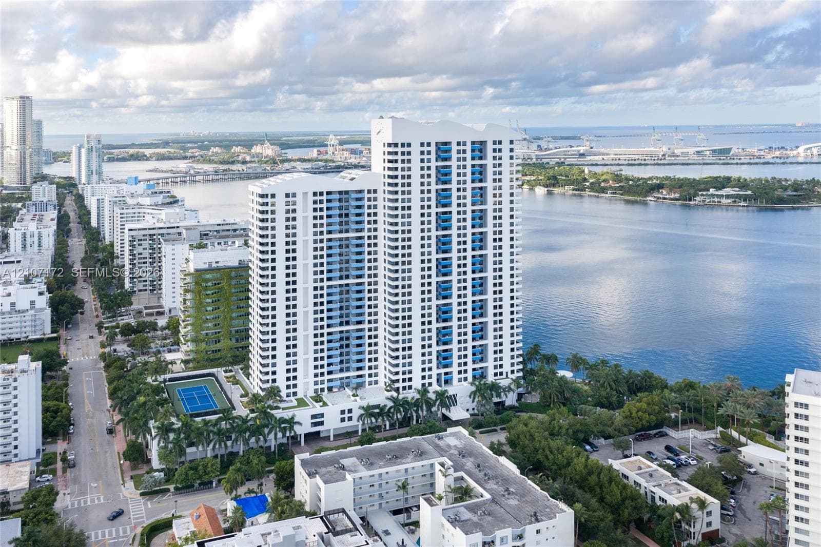 High-rise residential tower featuring white exterior cladding, recessed balconies, and an on-site tennis court, situated directly along the waterfront