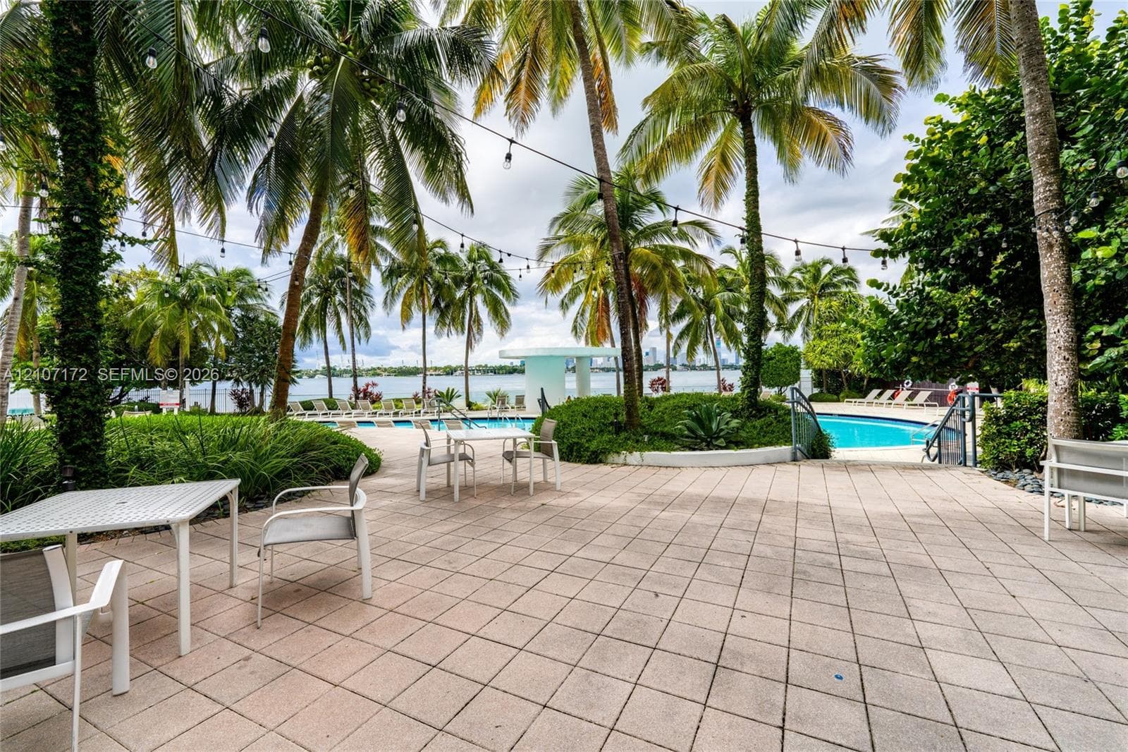 Outdoor patio featuring paved tile flooring, mature palm trees, string lighting, and an in-ground swimming pool overlooking the water