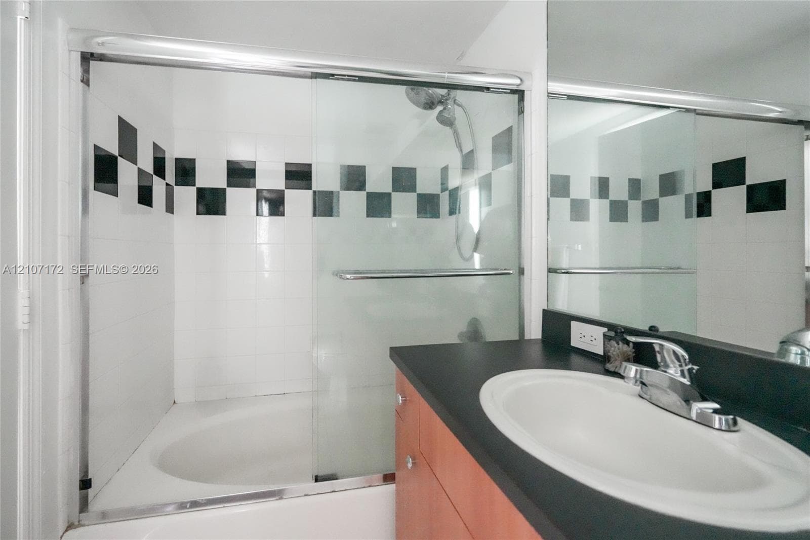 Bathroom featuring a tub-shower combination with glass sliding doors and a decorative black-and-white checkered tile accent, paired with a vanity containing a dark countertop, integrated oval basin, and chrome faucet