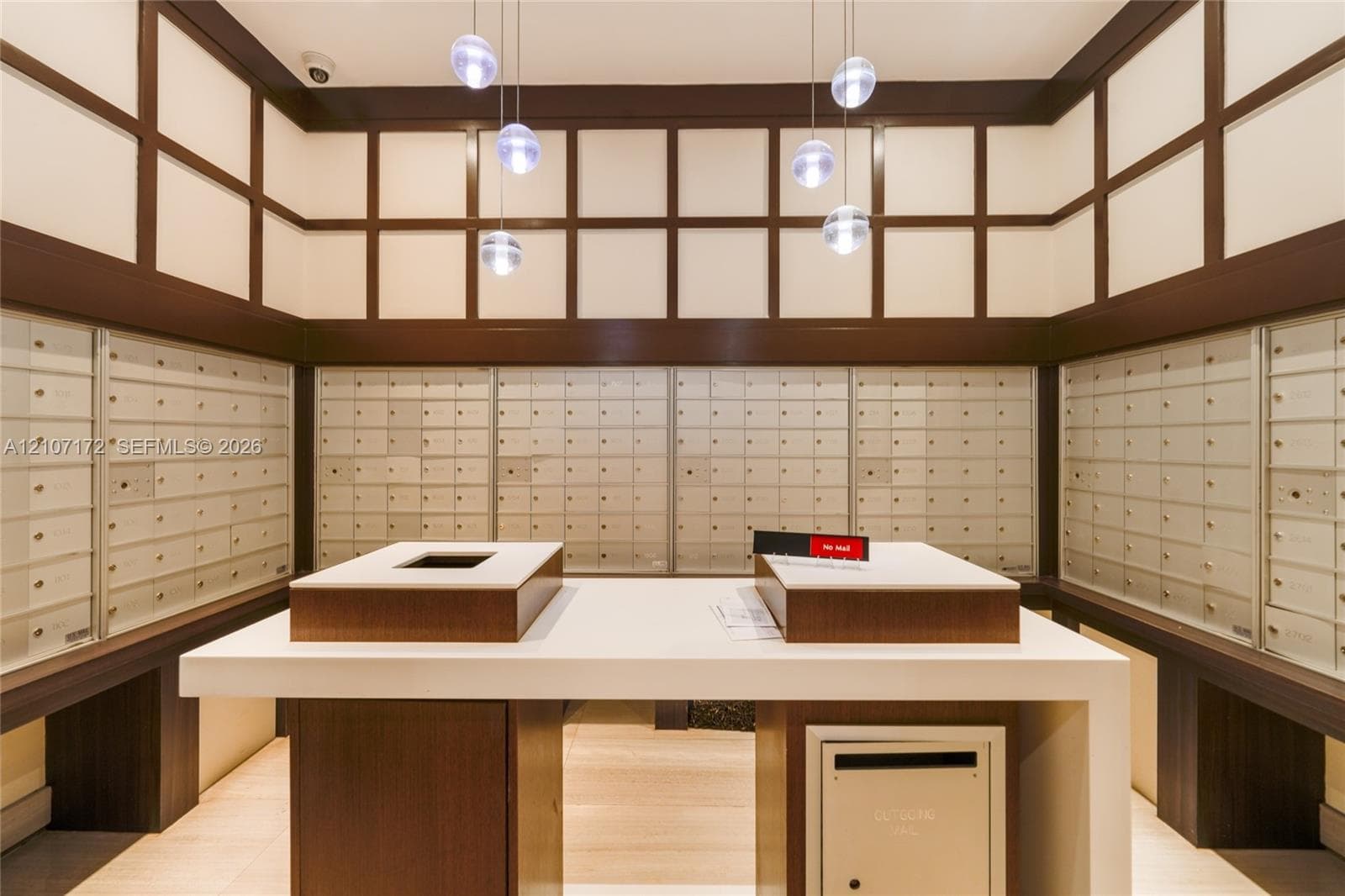 Mail room featuring integrated banks of metal parcel lockers, a central island with a solid surface countertop, wood-toned paneled walls with grid detailing, and suspended orb pendant lighting