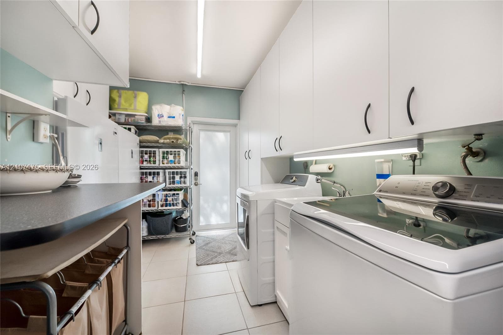 Laundry room featuring laundry tub, extensive white cabinetry, light-toned tile flooring, under-cabinet lighting, and impact glass exterior door.