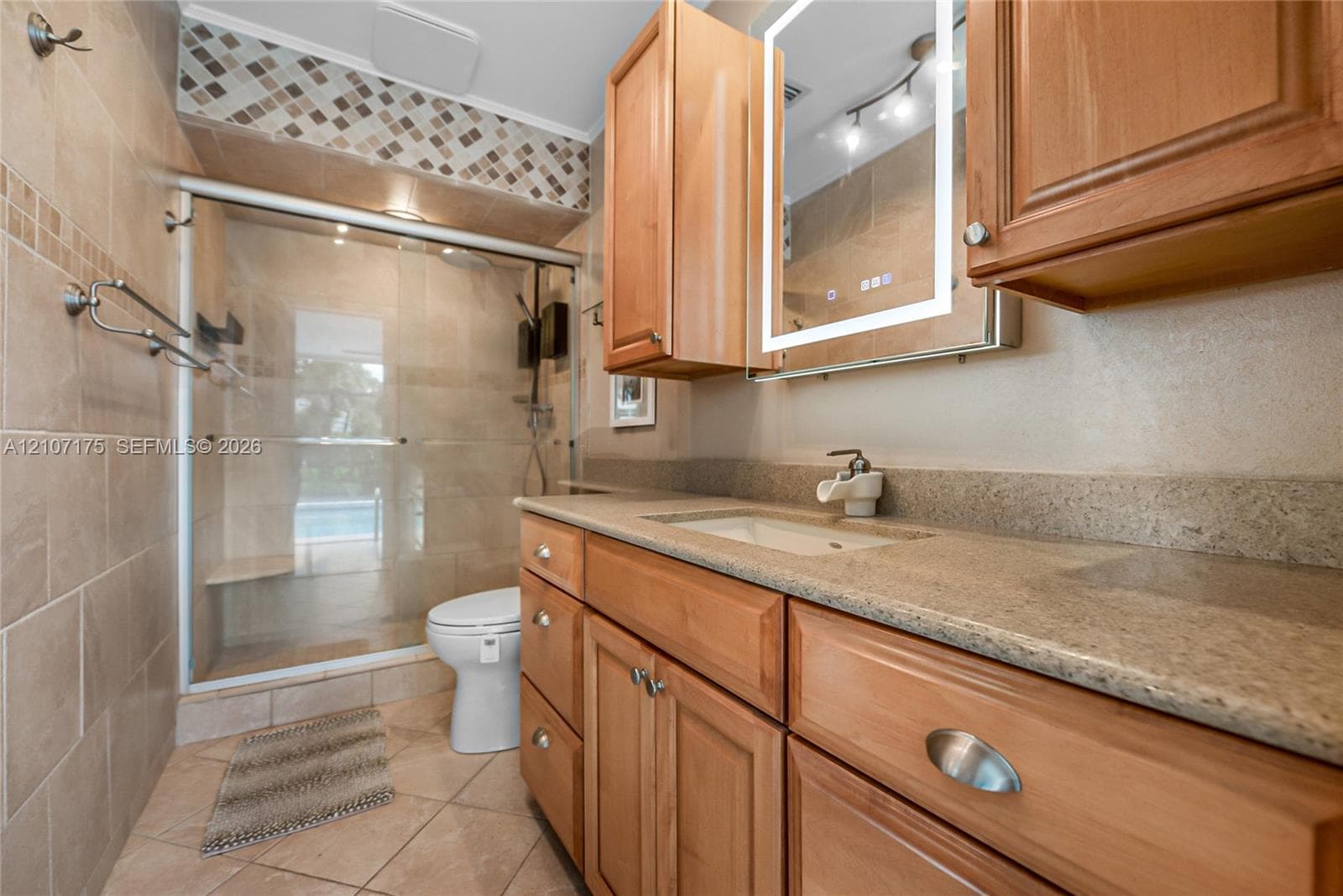 Bathroom featuring a wood-tone vanity with illuminated wall-mounted mirrored cabinet, walk-in shower with glass enclosure, and ceramic tile flooring