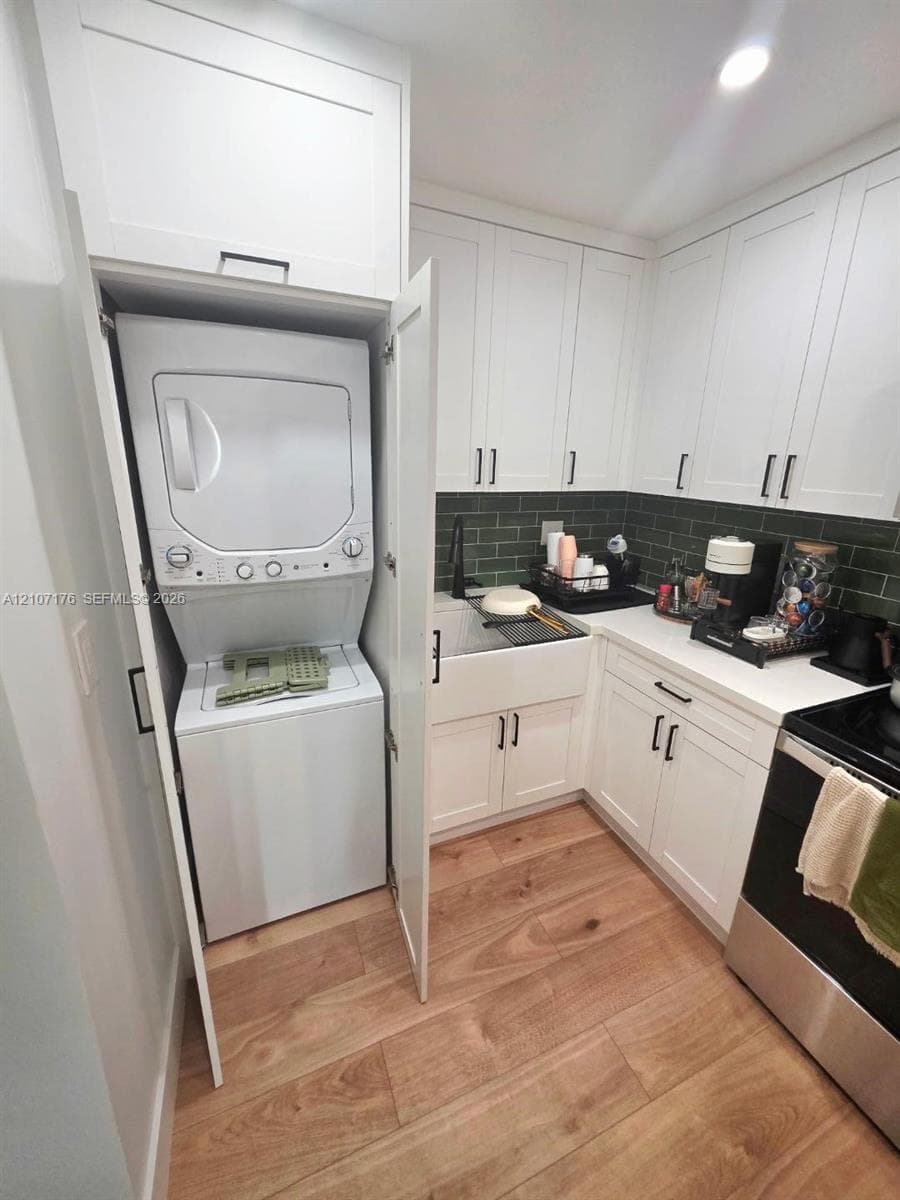 Shaker-style white cabinetry, farmhouse apron-front sink, green subway tile backsplash, wood-finish flooring, and built-in stacked laundry center