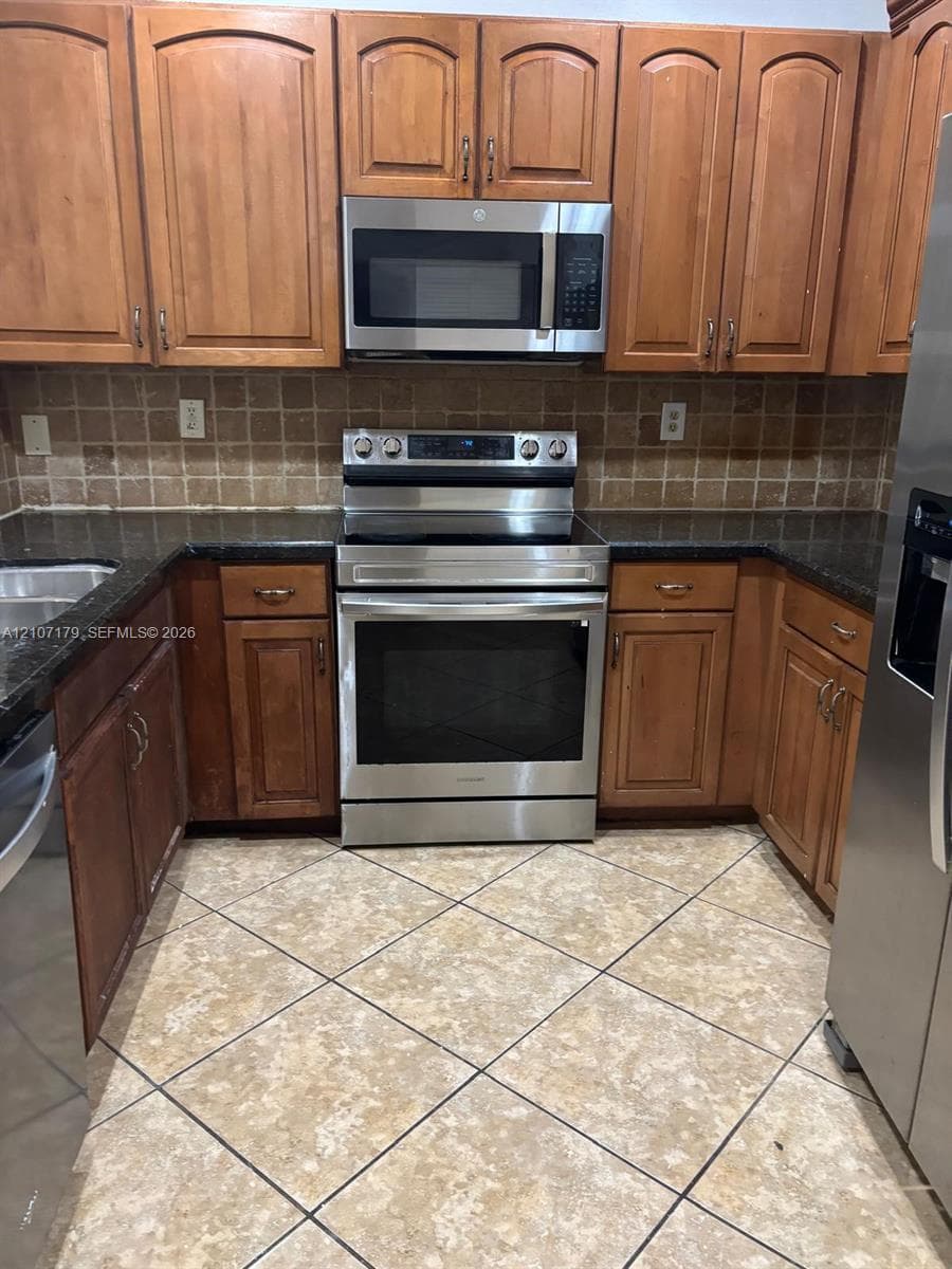 Kitchen featuring wood cabinetry, stone-finish countertops, tile backsplash, stainless steel appliances, and diagonal-set tile flooring