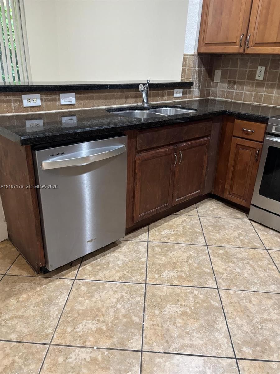 Kitchen featuring dark stone countertops, stainless steel dishwasher, wood-finish cabinetry, tile backsplash, and tile flooring