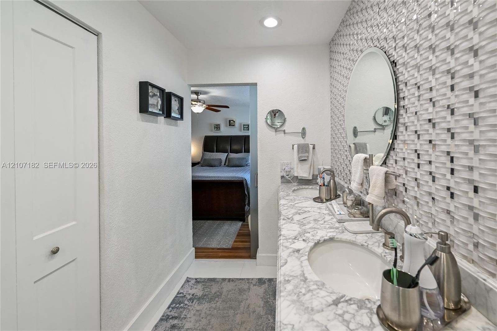 Dual-sink vanity with patterned stone countertop, custom mosaic tile backsplash, recessed lighting, and oval wall-mounted mirrors
