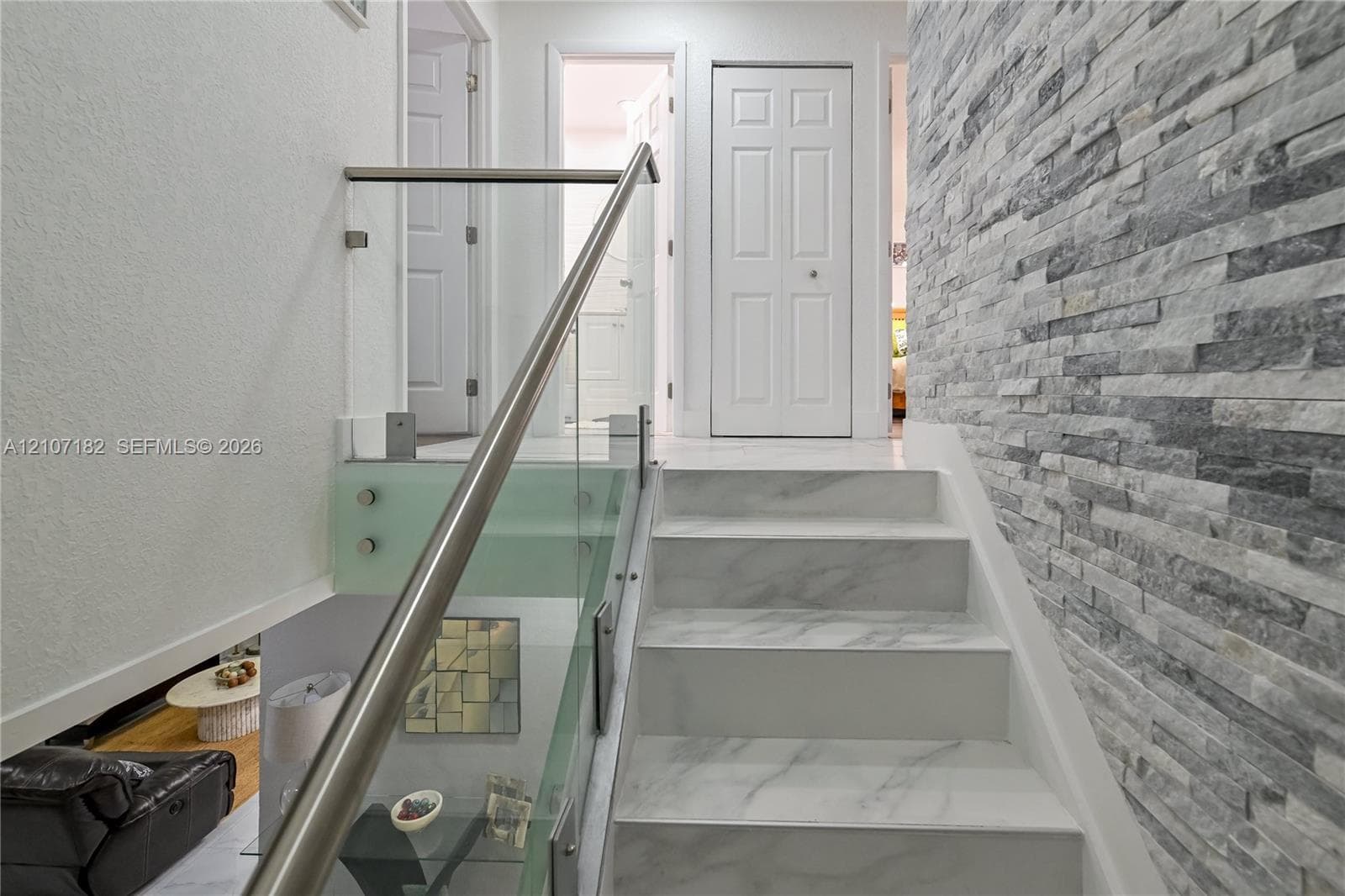 Staircase featuring white stone-finish treads, a stacked-stone accent wall, and a glass panel railing with a metallic handrail