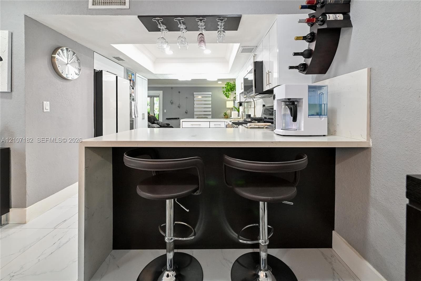 Waterfall peninsula featuring a solid-surface countertop and contrasting dark cabinetry, overhead wine glass rack, tray ceiling, and white floor tiling with veining