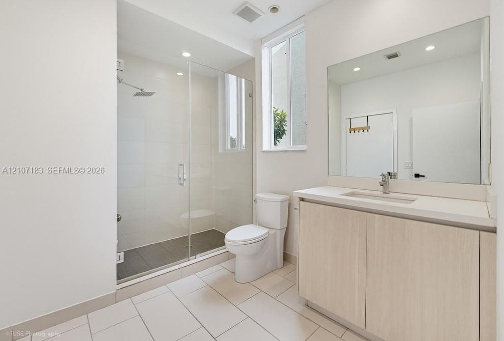 Bathroom featuring a glass-enclosed walk-in shower with a rain-style showerhead, wood-finish vanity with a rectangular basin, light-toned tile flooring, and a large wall-mounted mirror