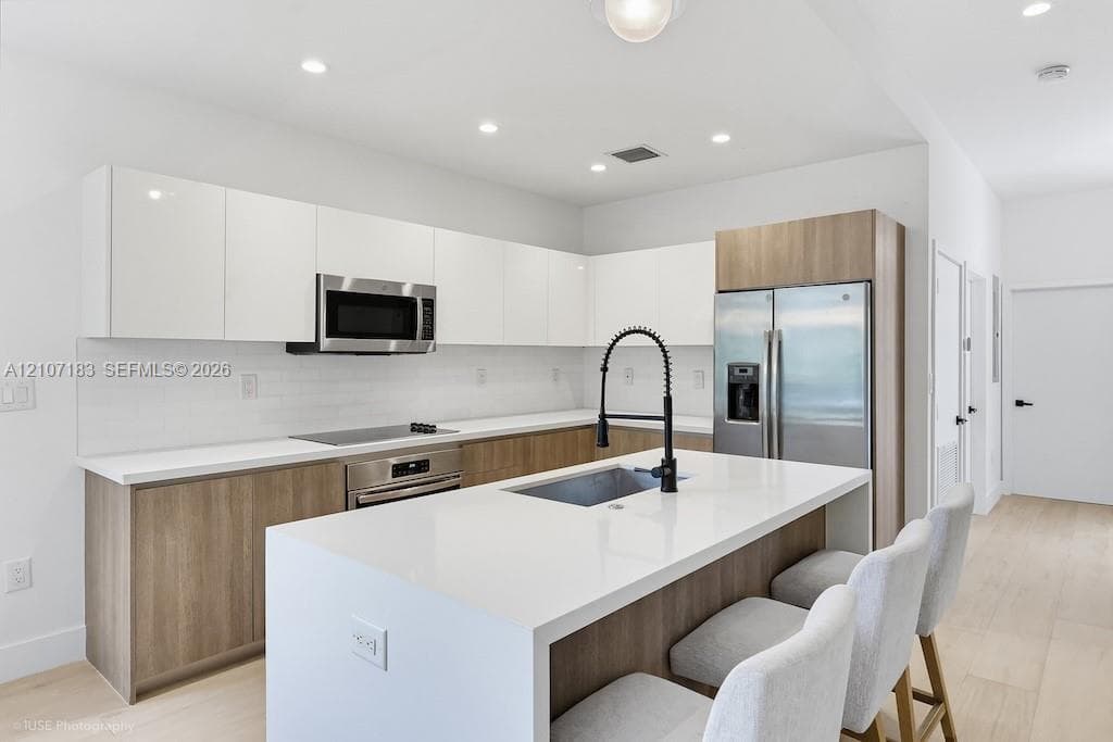 Kitchen featuring flat-panel two-tone cabinetry, stone-surface countertops, a center island with an integrated sink and gooseneck faucet, stainless steel appliances, and wood-finish flooring