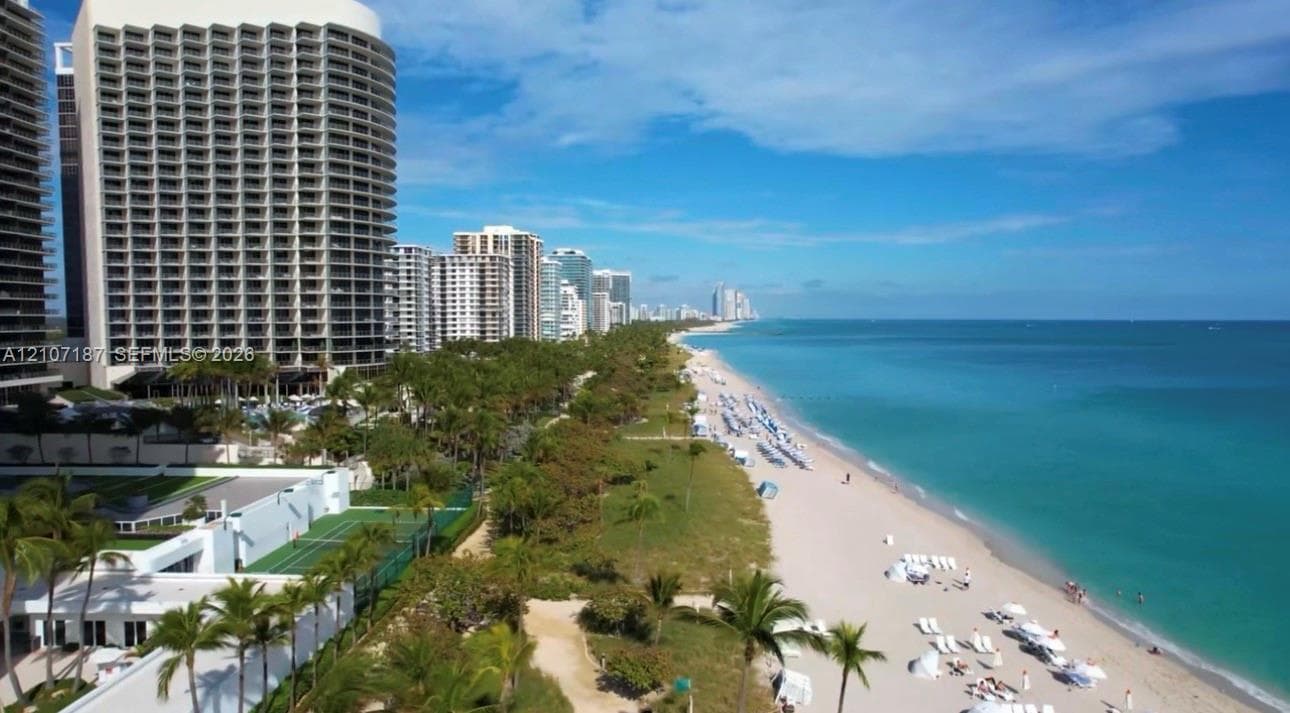 Coastal high-rise building with direct oceanfront access, featuring a private tennis court and lush palm-lined grounds situated along the sandy shoreline