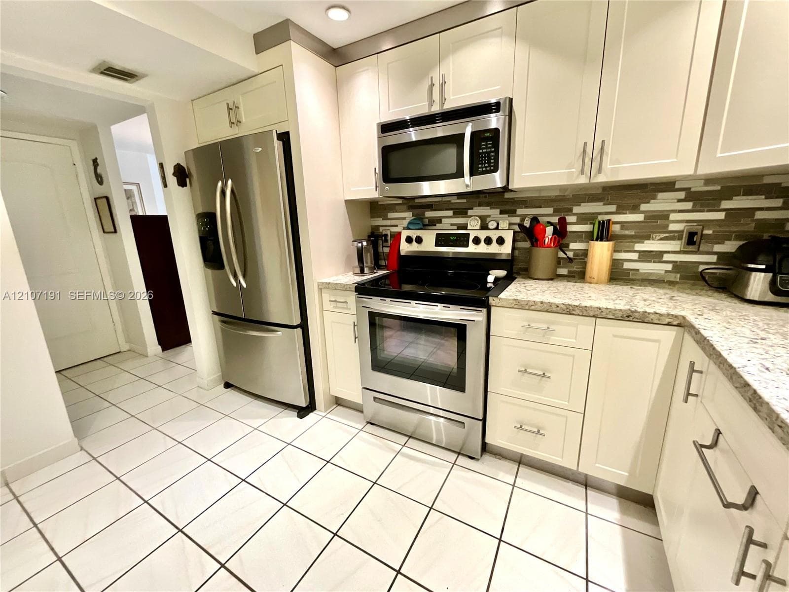 Kitchen featuring shaker-style cabinetry, stainless steel appliances, multi-tonal linear tile backsplash, patterned stone countertops, and tile flooring