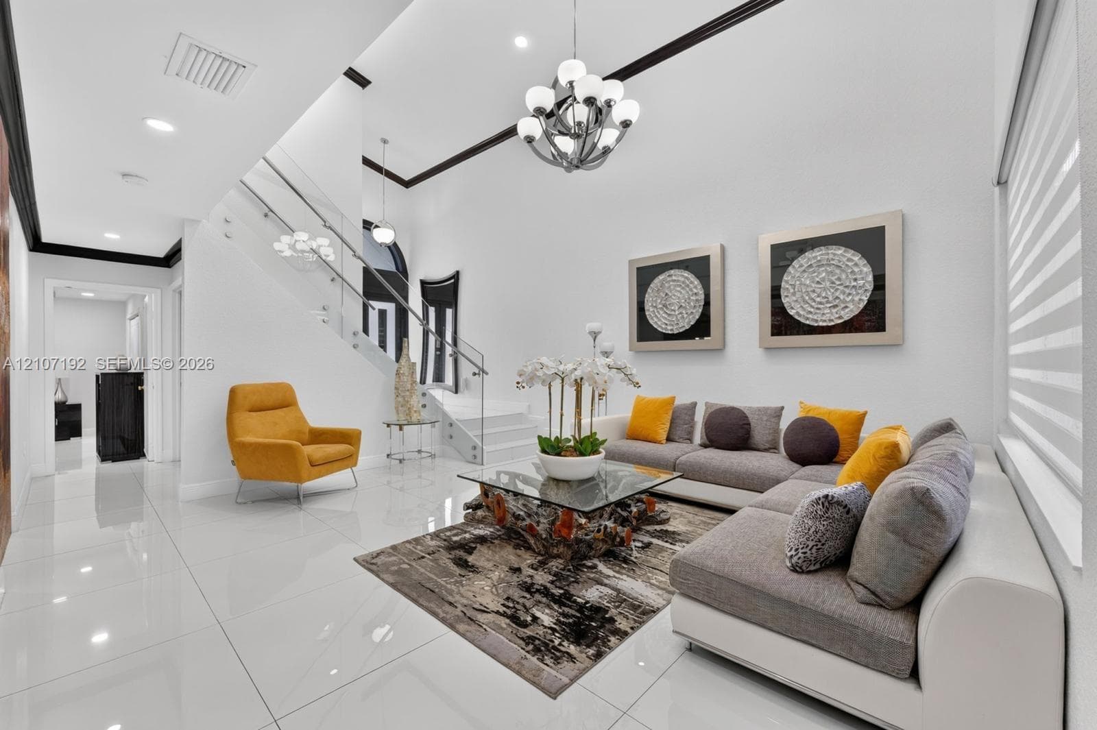 Open-concept interior featuring large-format polished tile flooring, a glass-panel staircase railing, high ceilings with crown molding, and an integrated multi-tiered chandelier