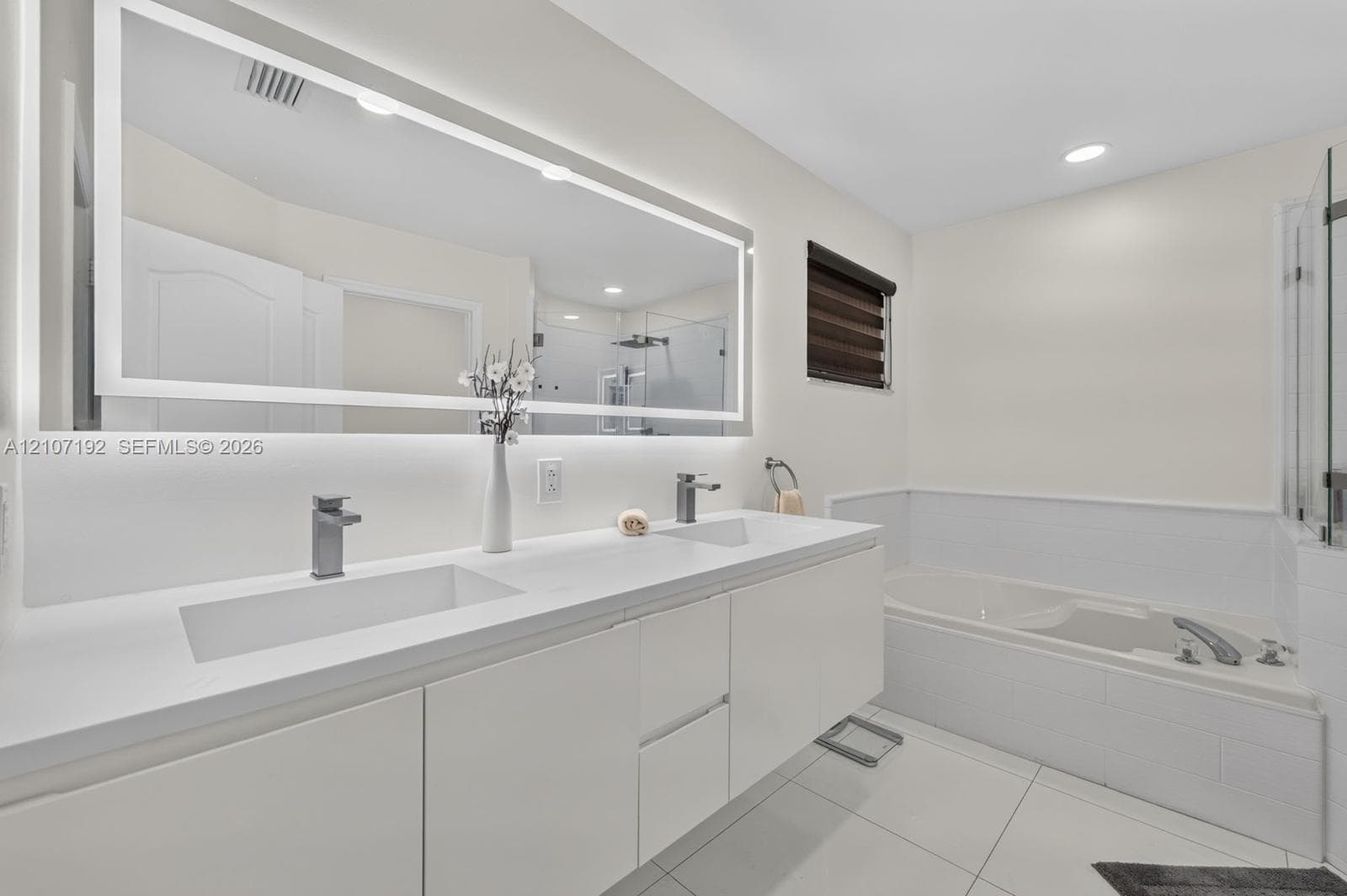 Dual-sink vanity with white cabinetry, integrated stone-surface countertop, full-width LED-backlit mirror, and separate soaking tub with glass-enclosed shower surround