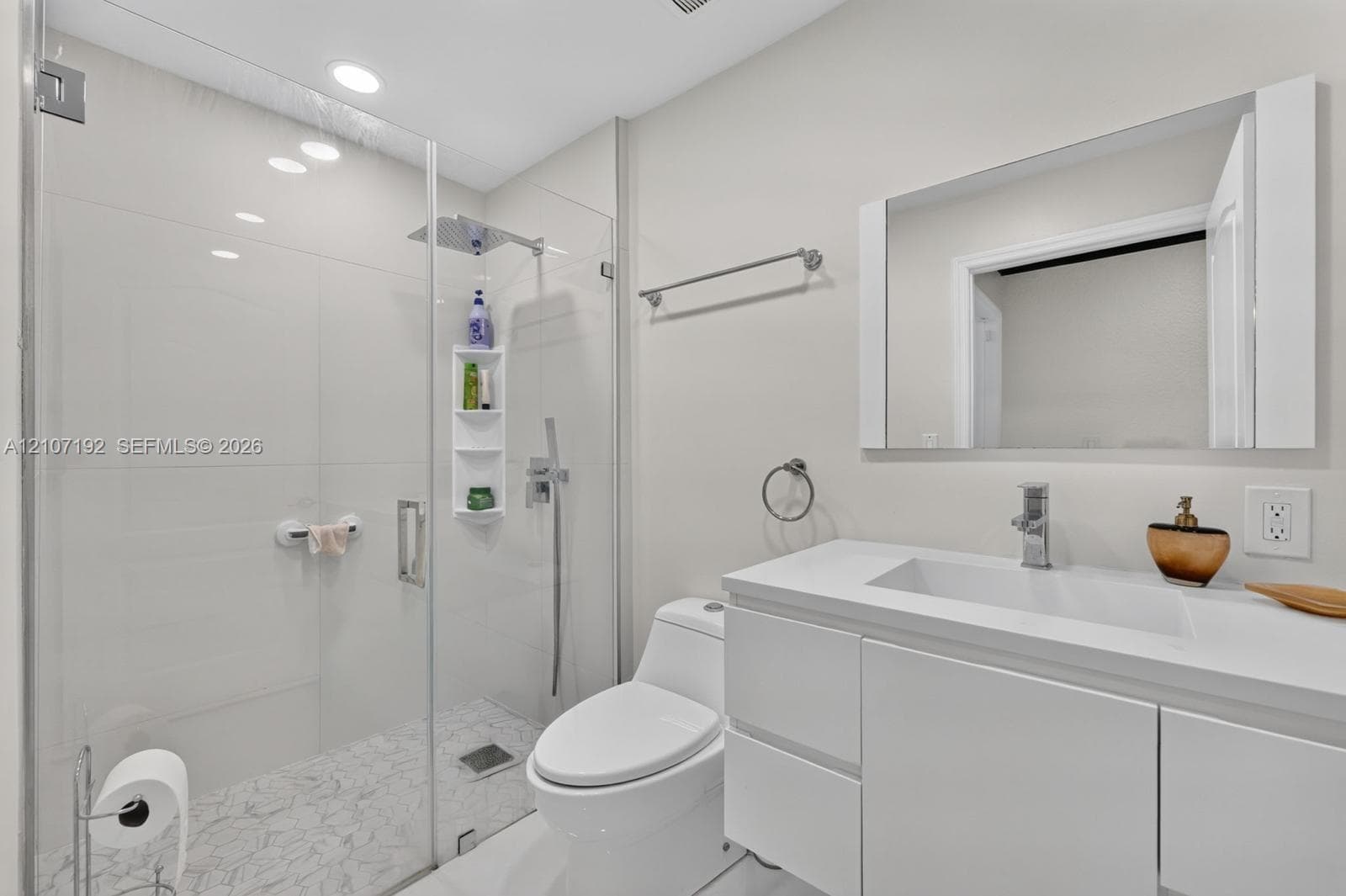 Modern bathroom featuring a frameless glass shower enclosure, rainfall shower head with handheld wand, integrated storage niche, sleek white vanity with a rectangular sink, and recessed lighting